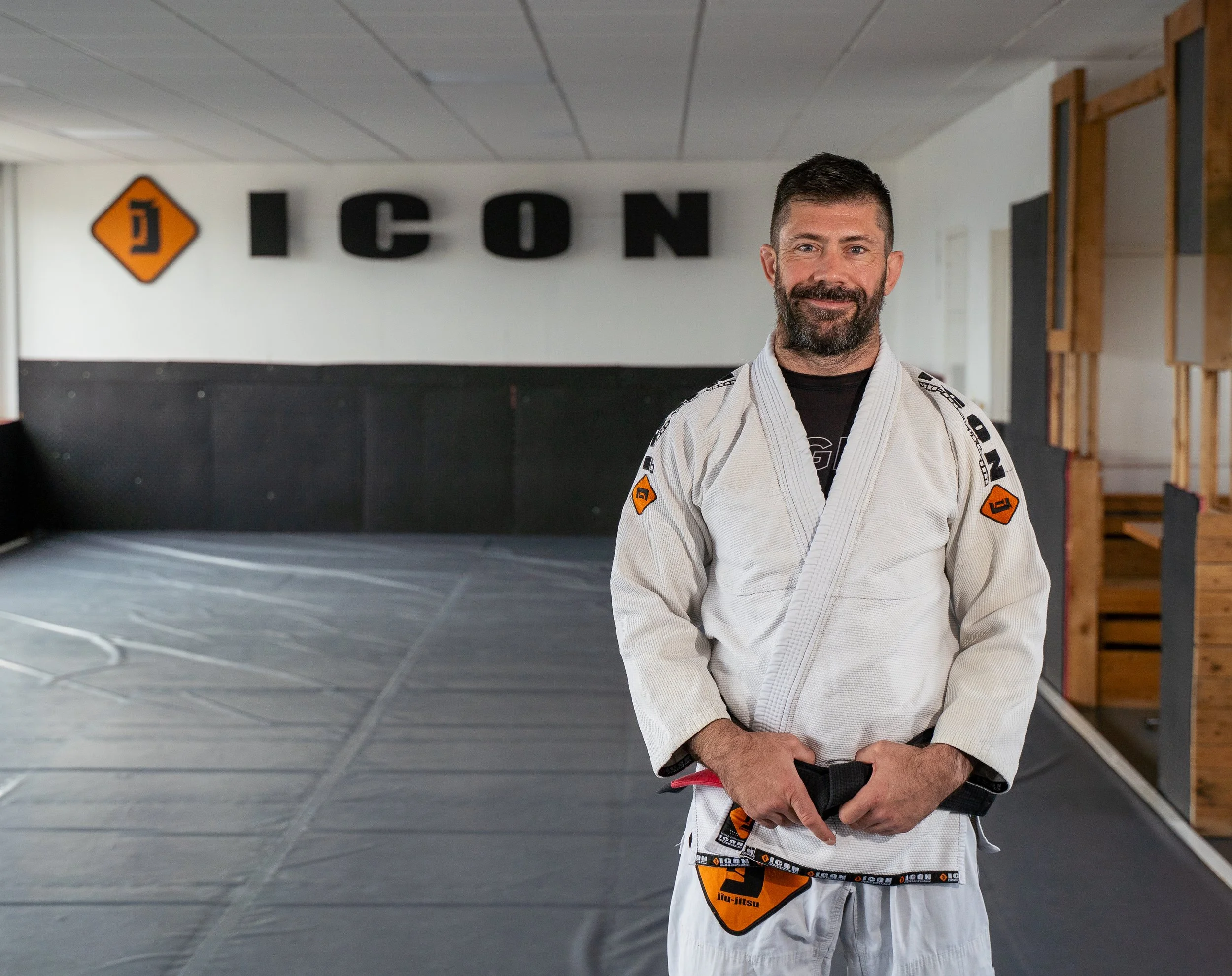 A man with a beard in a white Brazilian Jiu-Jitsu gi standing in a martial arts gym, smiling at the camera, with the word 'ICON' on the wall behind him.