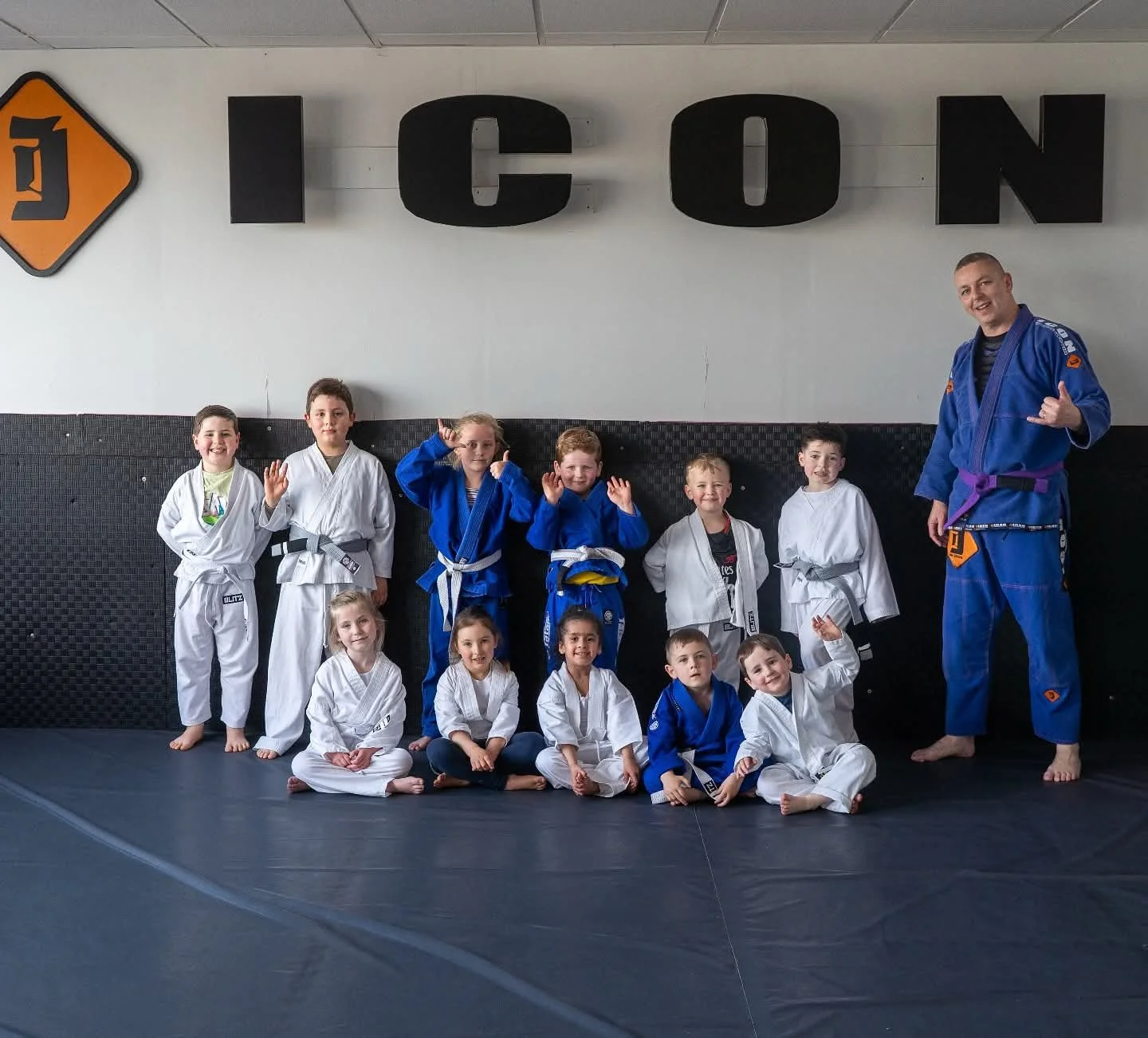 Children in martial arts uniforms with an instructor posing in a dojo, some children are sitting on the floor, others standing, illustrating a group class or recognition event.