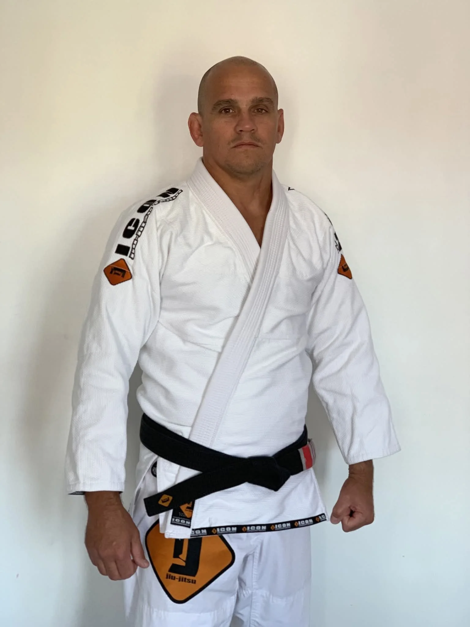 An older man in a white Brazilian Jiu-Jitsu gi with black belt, standing against a plain white wall, looking serious.