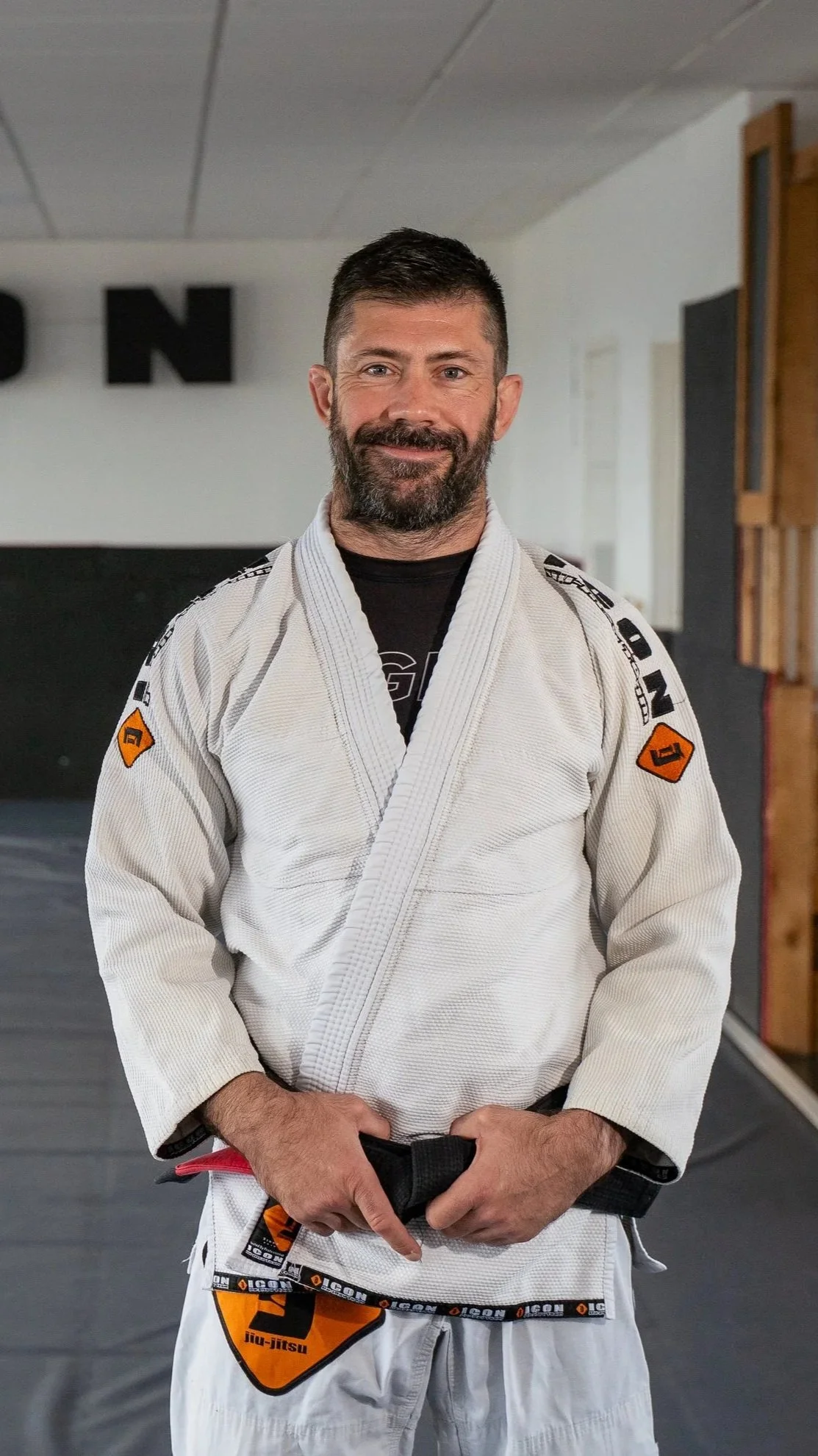A man with a beard in a Brazilian Jiu-Jitsu gi standing in a martial arts gym or dojo.