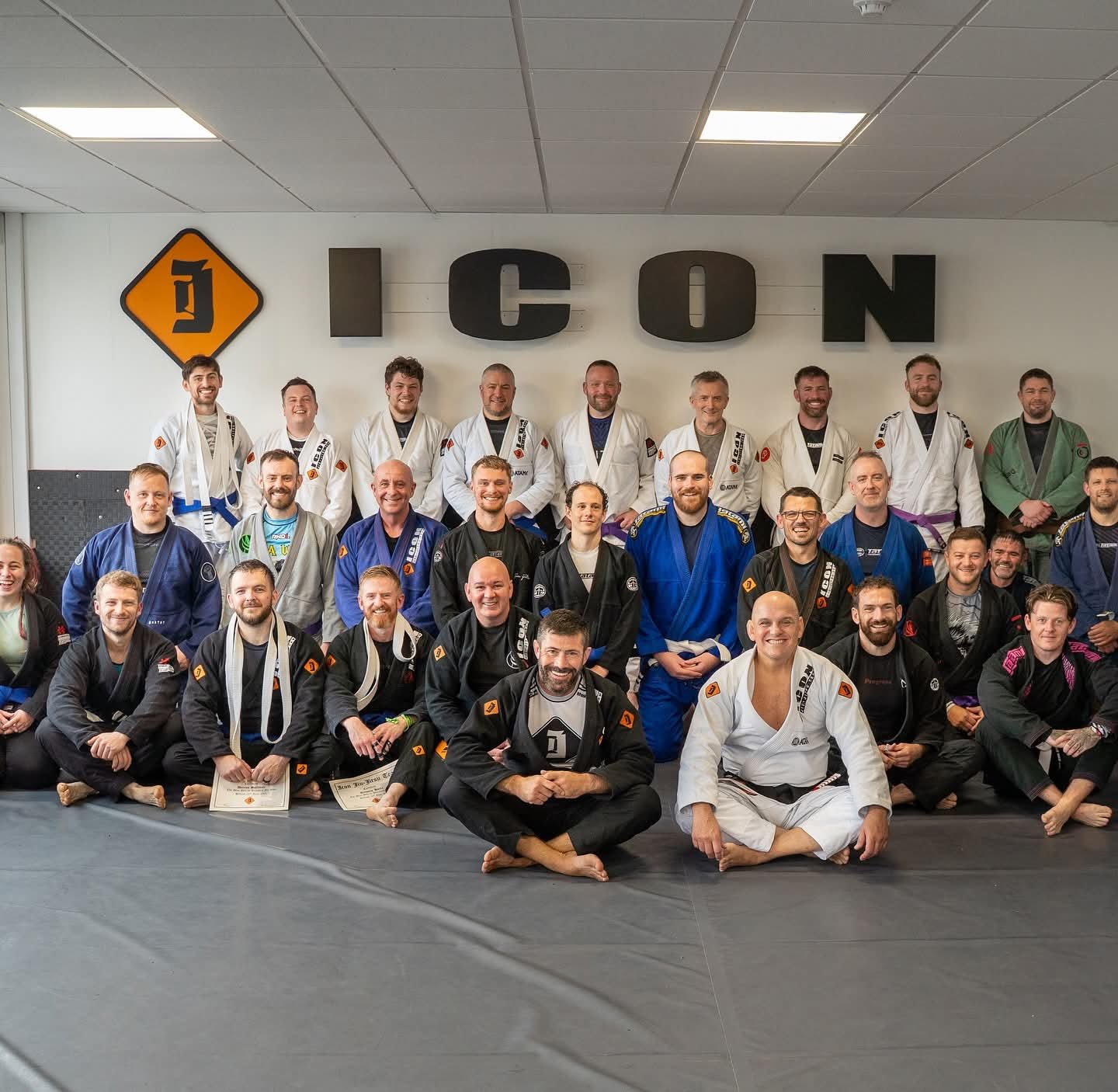 Group of Brazilian Jiu-Jitsu practitioners in gis and belts posing for a photo in a training facility with a sign that reads 'ICON' on the wall.