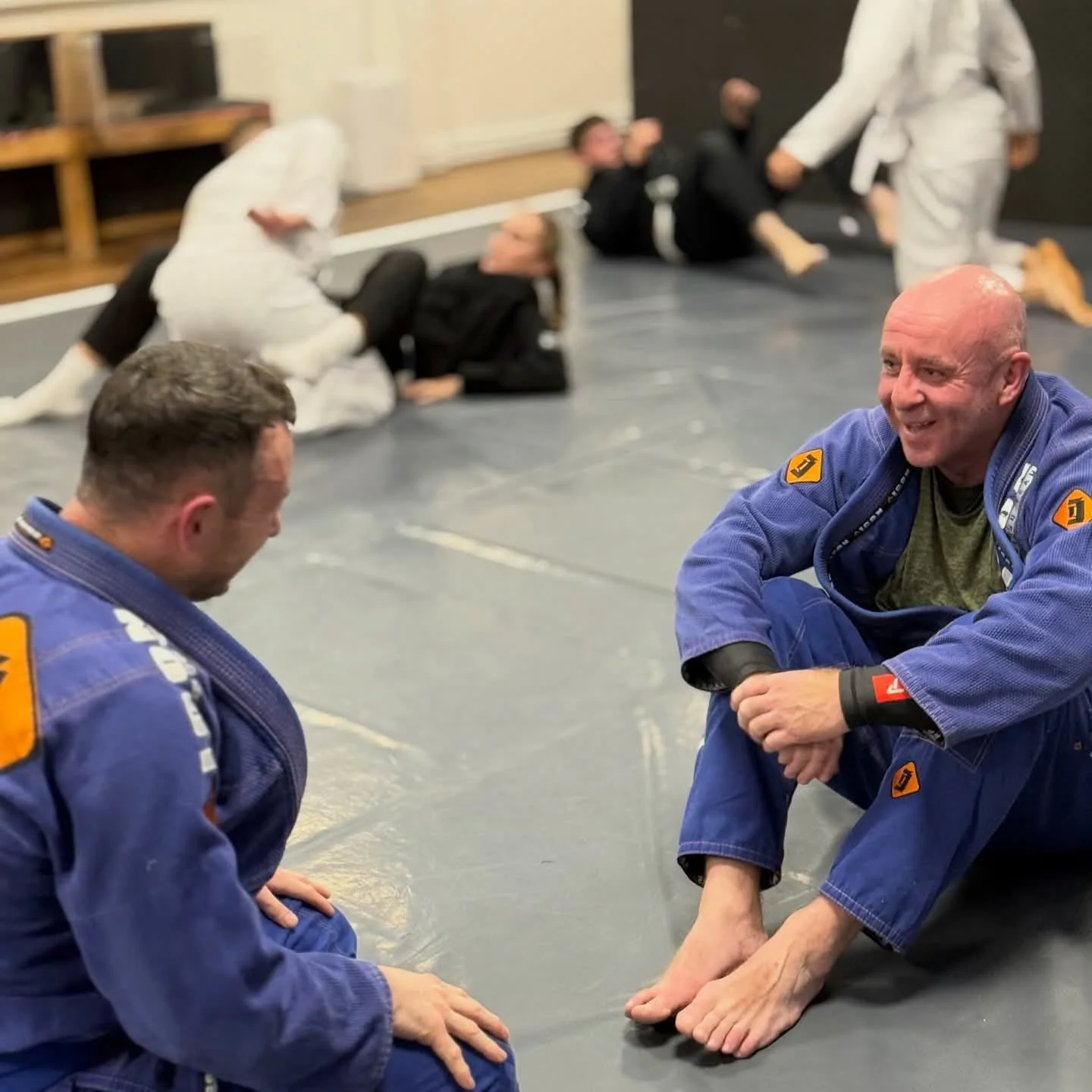 Two Brazilian jiu-jitsu practitioners practicing on a mat, with four more practitioners lying or sitting in the background, one of whom is being attended to by an instructor.