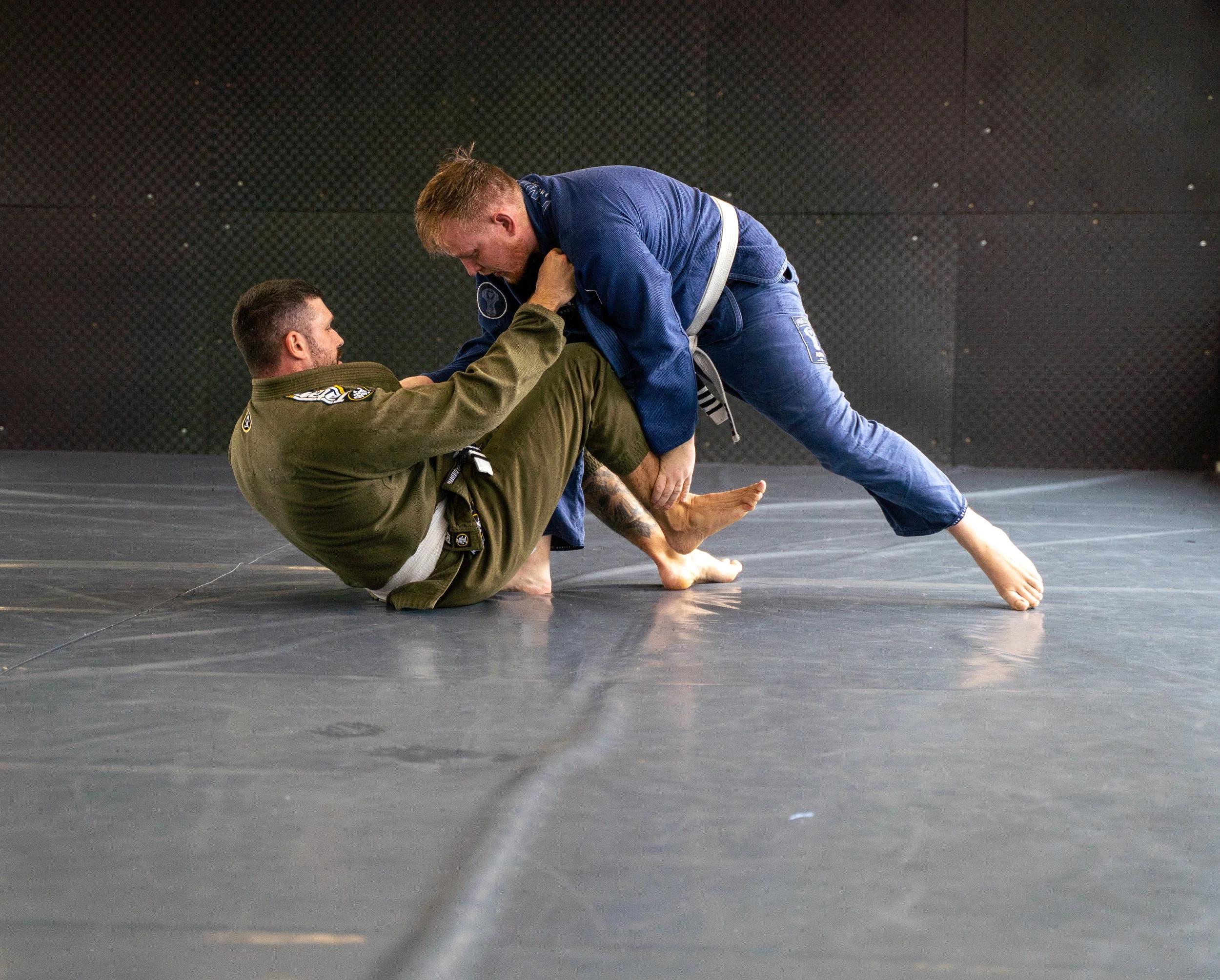 Two men demonstrating Brazilian Jiu-Jitsu on a training mat, one in a green gi and the other in a blue gi, engaging in a ground fighting position.