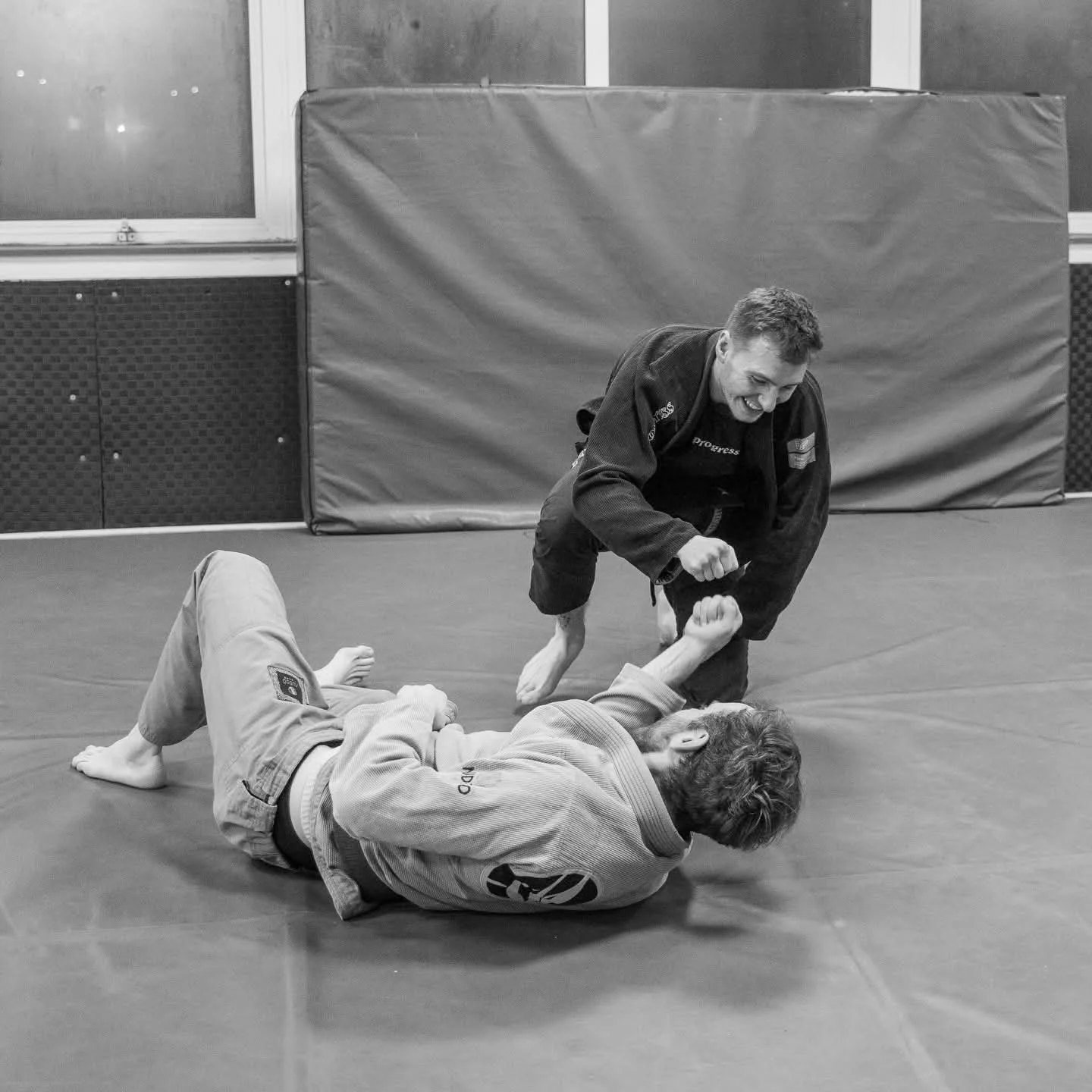 Two men practicing Brazilian Jiu-Jitsu in a gym, with one on the ground and the other standing, both wearing gis, on a padded mat.