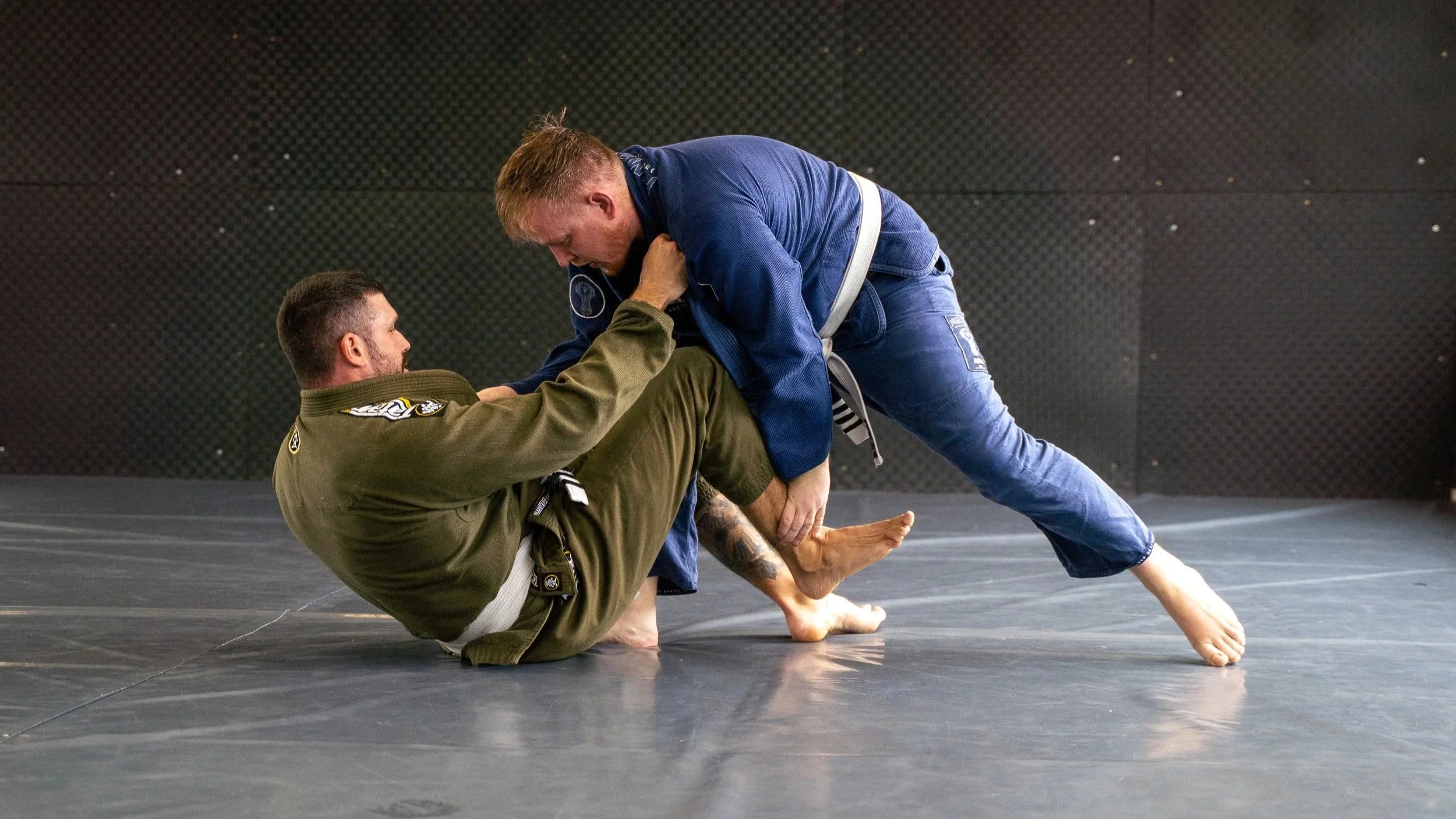 Two men practicing Brazilian Jiu-Jitsu on a mat, wearing gis, one in green and the other in blue, engaged in a grappling position.