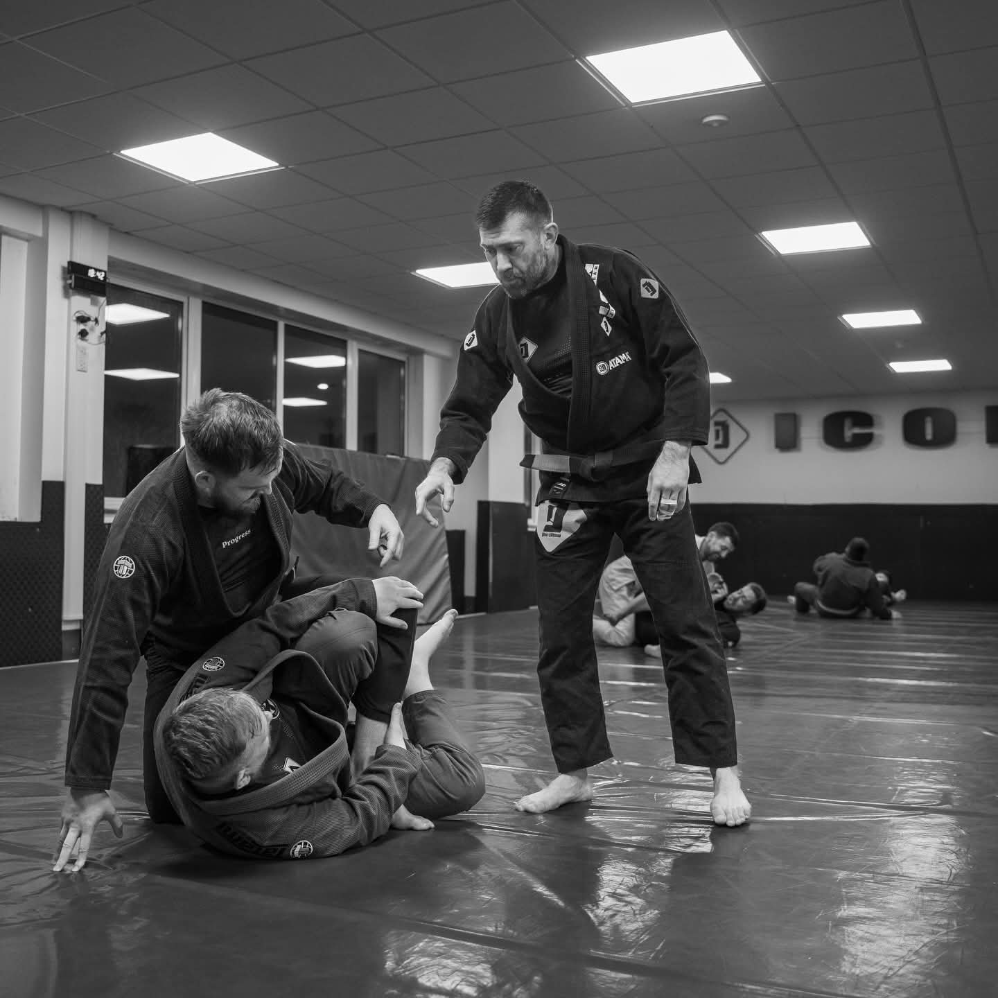 Two male Brazilian Jiu-Jitsu practitioners in gis practicing a technique on a mat in a gym, with other practitioners training in the background.