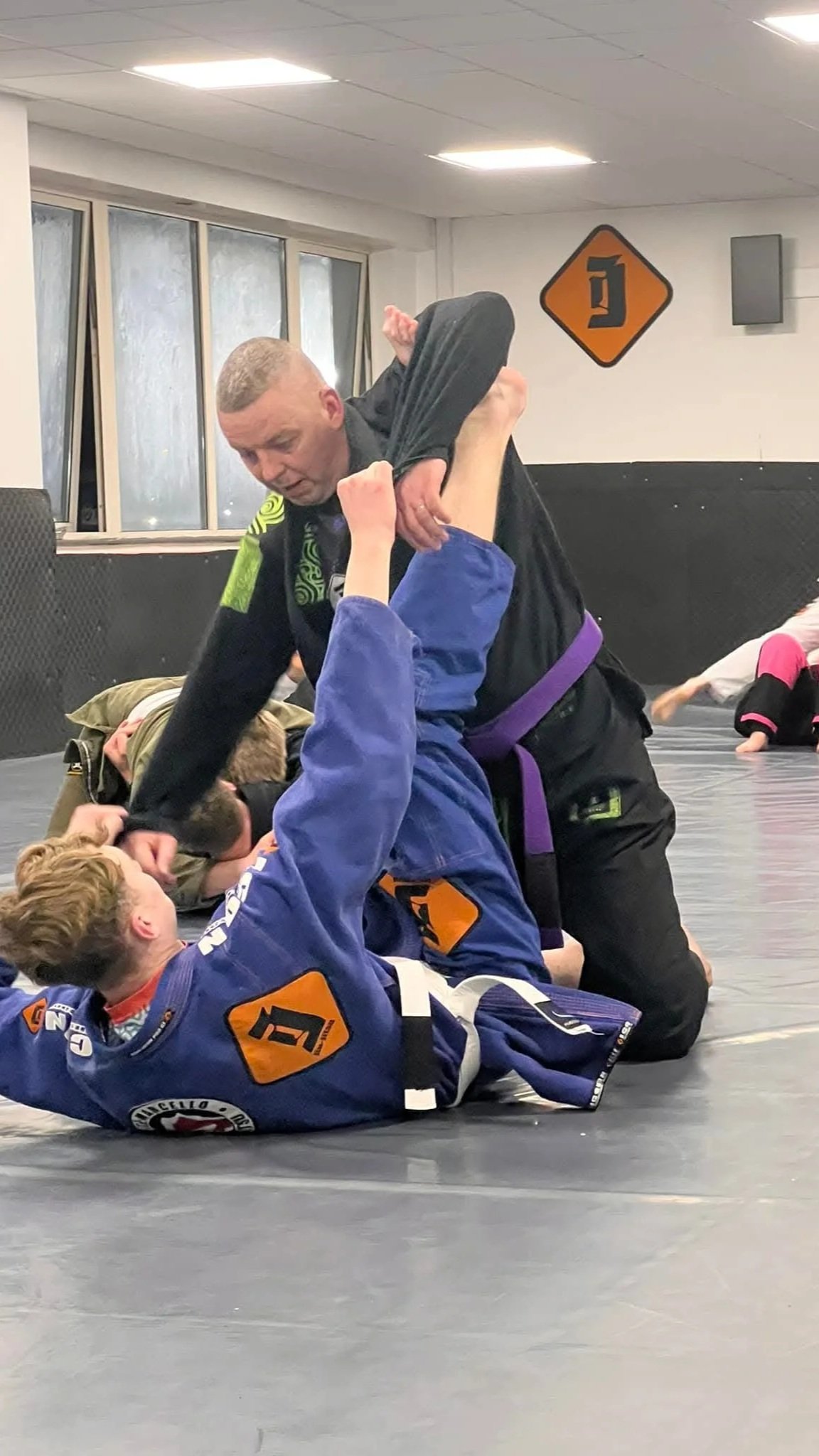 Two kids practicing Brazilian Jiu-Jitsu on a mat, one in a blue gi and the other in a green gi, with an instructor assisting. The instructor is wearing a black gi with a purple belt.