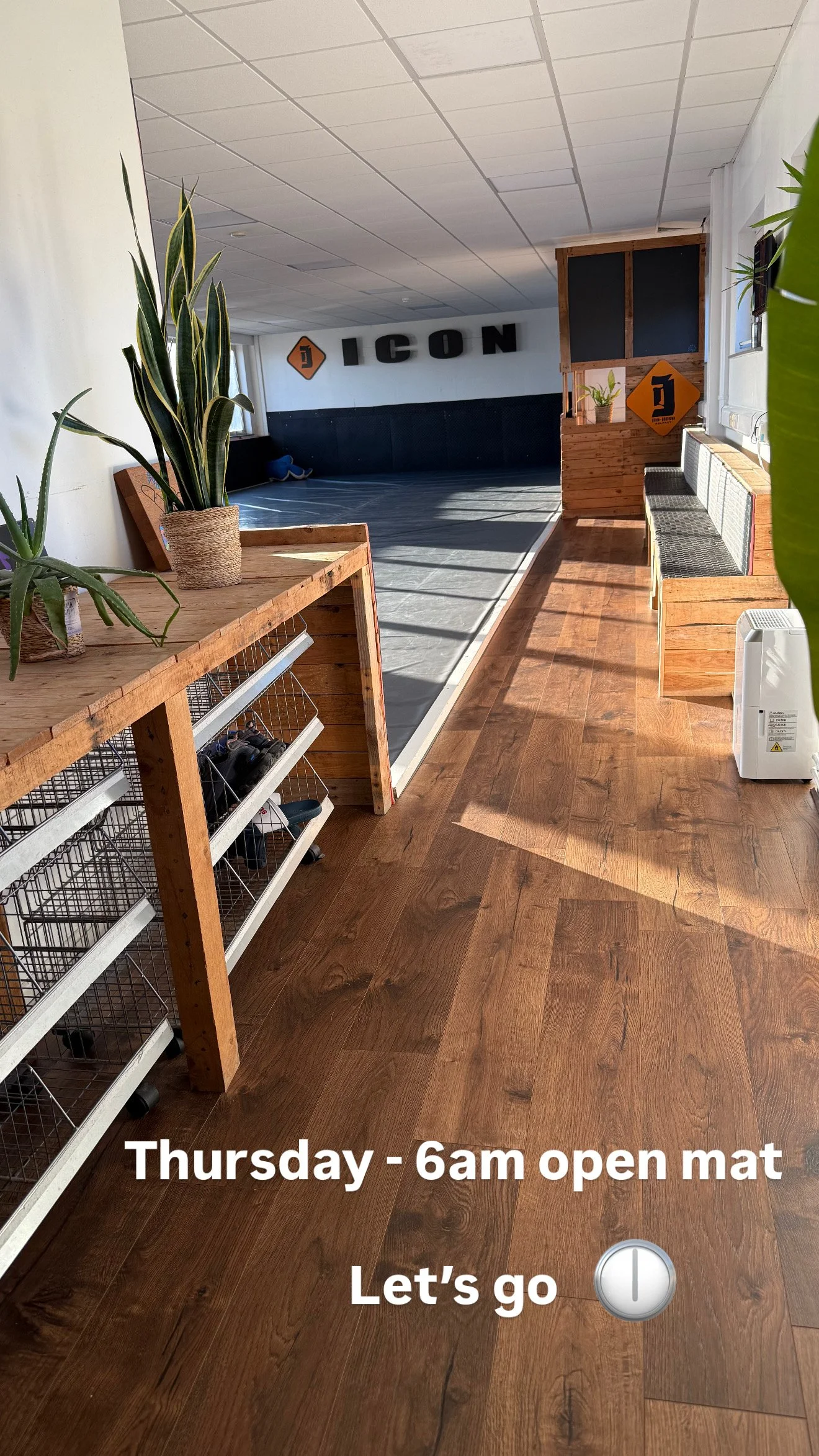 An interior room with wooden floors and decor, including potted plants and a black mat area. There are black and orange signs on the walls spelling 'ICON'. Text overlay indicates an event on Thursday at 6am, with a message 'Let's go' accompanied by a clock emoji.