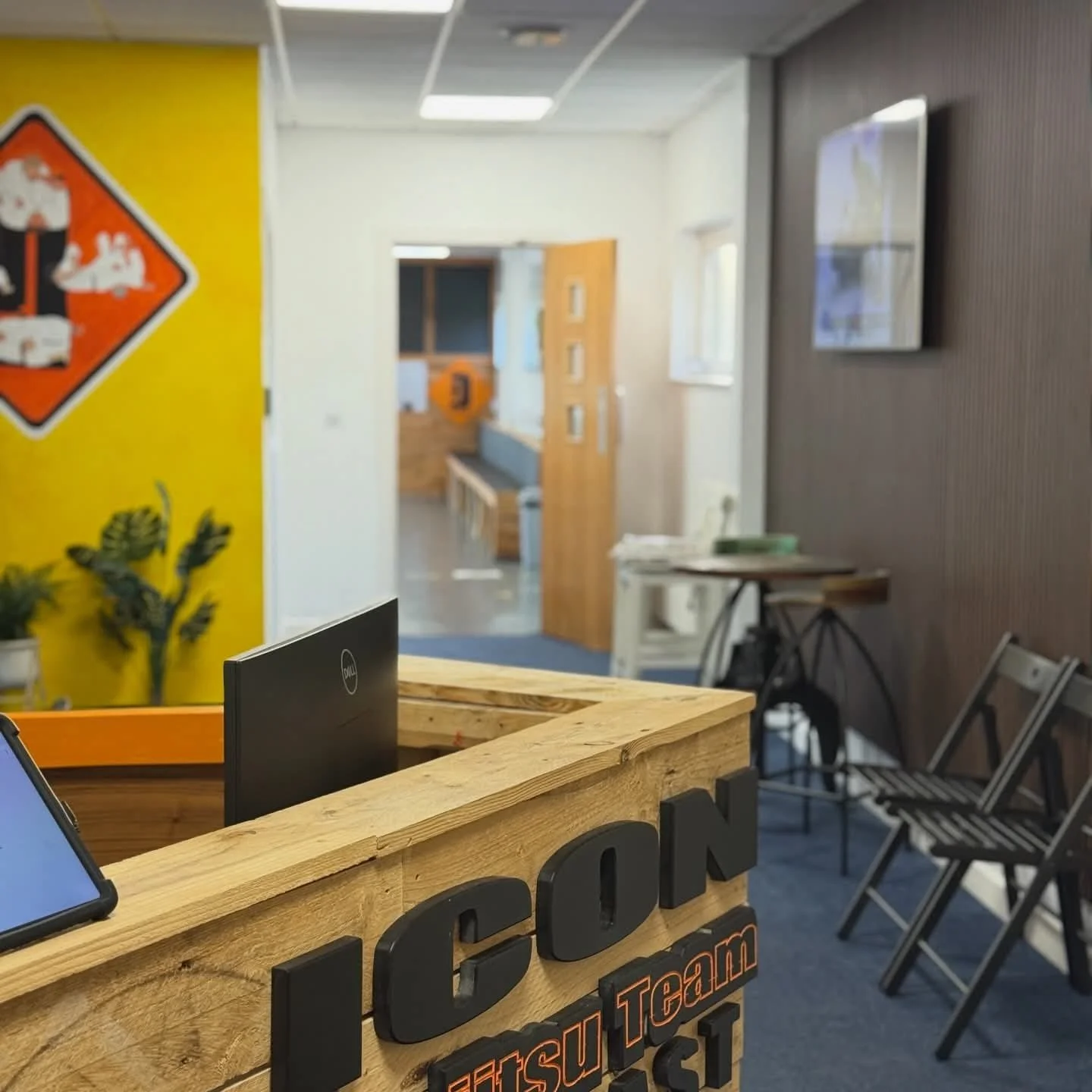 Indoor reception area with a wooden desk displaying the logo 'ICCON Futsu Team' in black and orange. Behind the desk, a blurred background features a sitting area, a plant on the left, and a yellow wall with a warning sign. There are chairs along the right wall, an open door leading to another room, and a wall-mounted TV on the right.