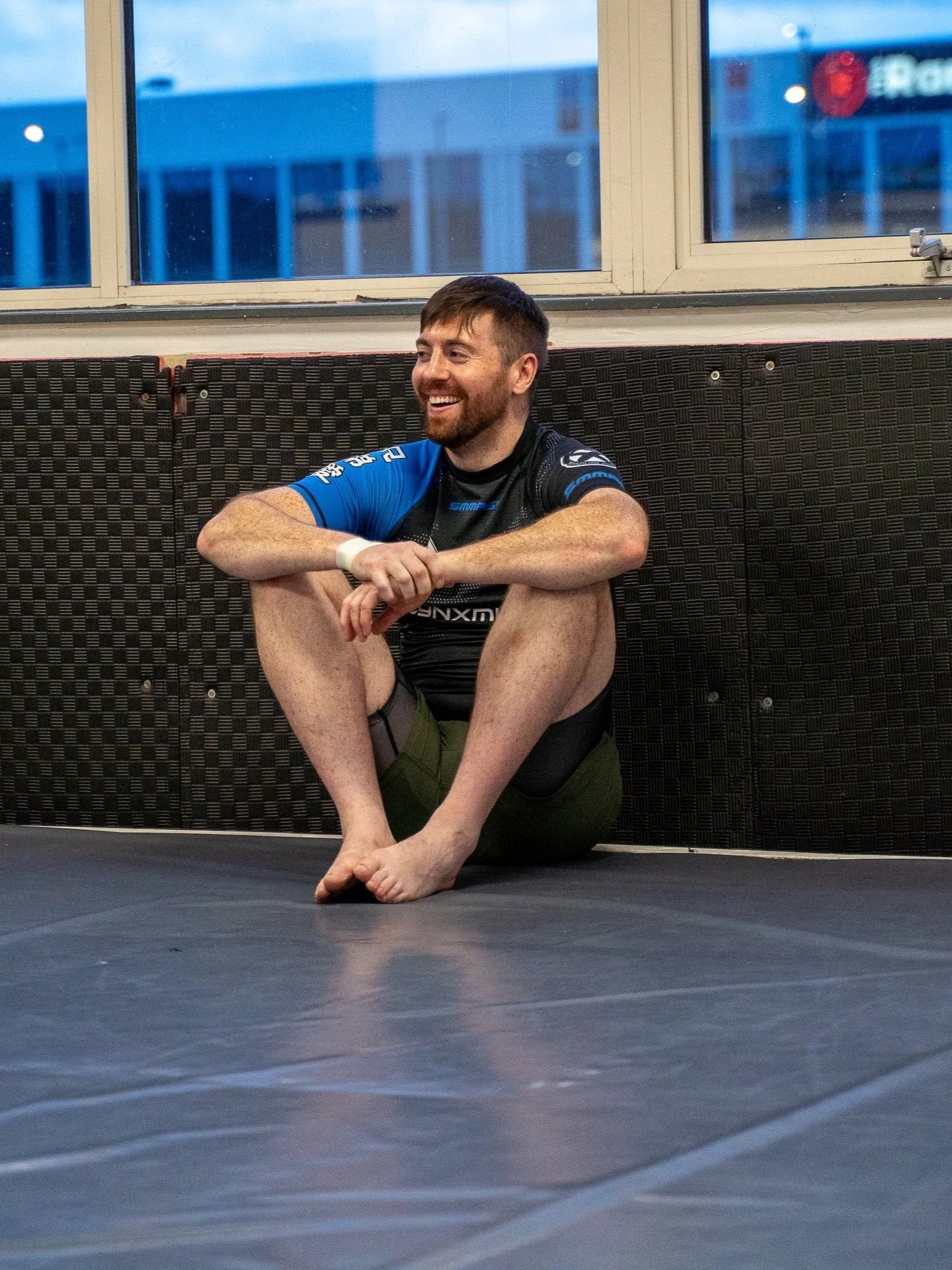 A man with brown hair and a beard sitting on the floor with his knees up, smiling and looking to the side, wearing athletic gear, in a gym near a window.