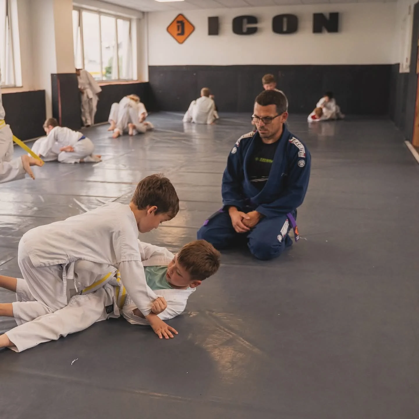 Children practicing Brazilian Jiu-Jitsu on mats with an instructor observing.