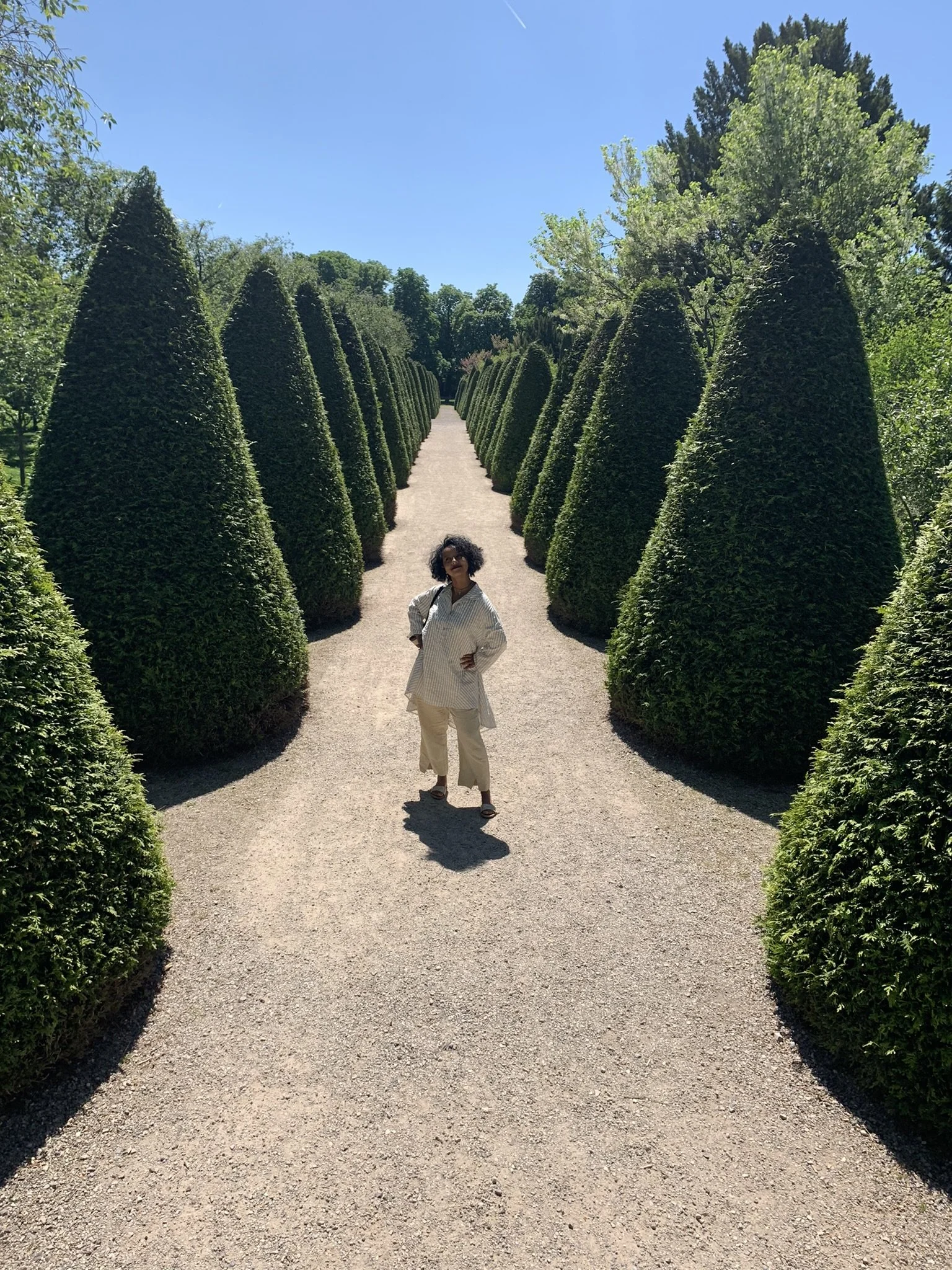 A woman standing in the middle of a gravel pathway flanked by tall, trimmed, cone-shaped bushes in a garden or park on a sunny day.
