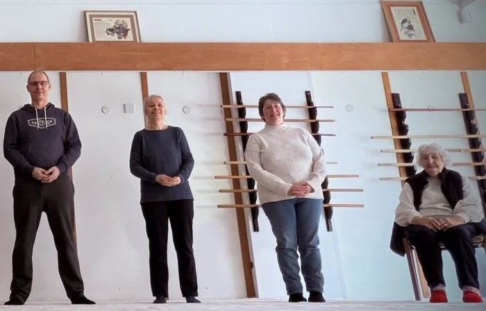 Four Qigong participants stand and sit in a room with wooden wall bars and framed pictures on the wall.