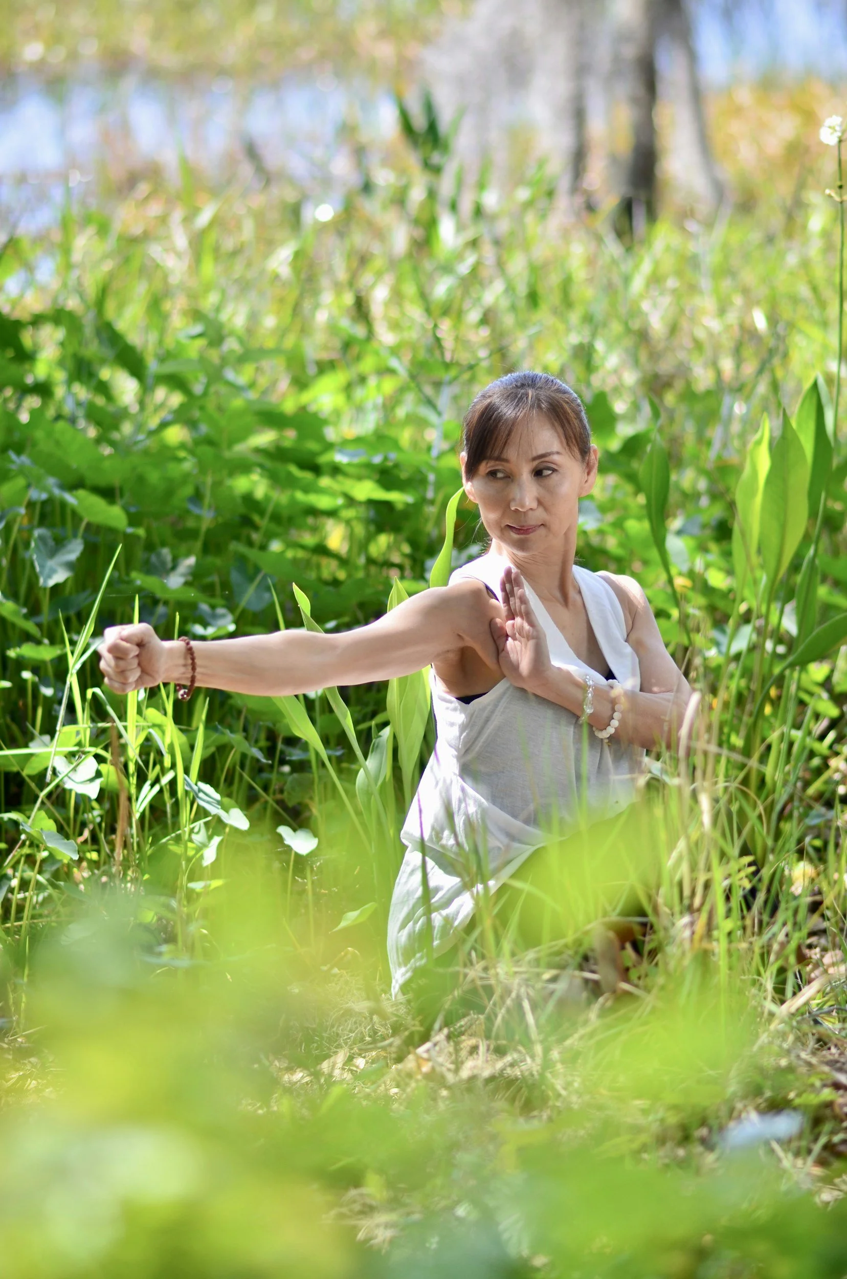 A woman practicing yoga outdoors in a lush green forest, performing a side stretch with one arm extended and the other arm bent across her chest.