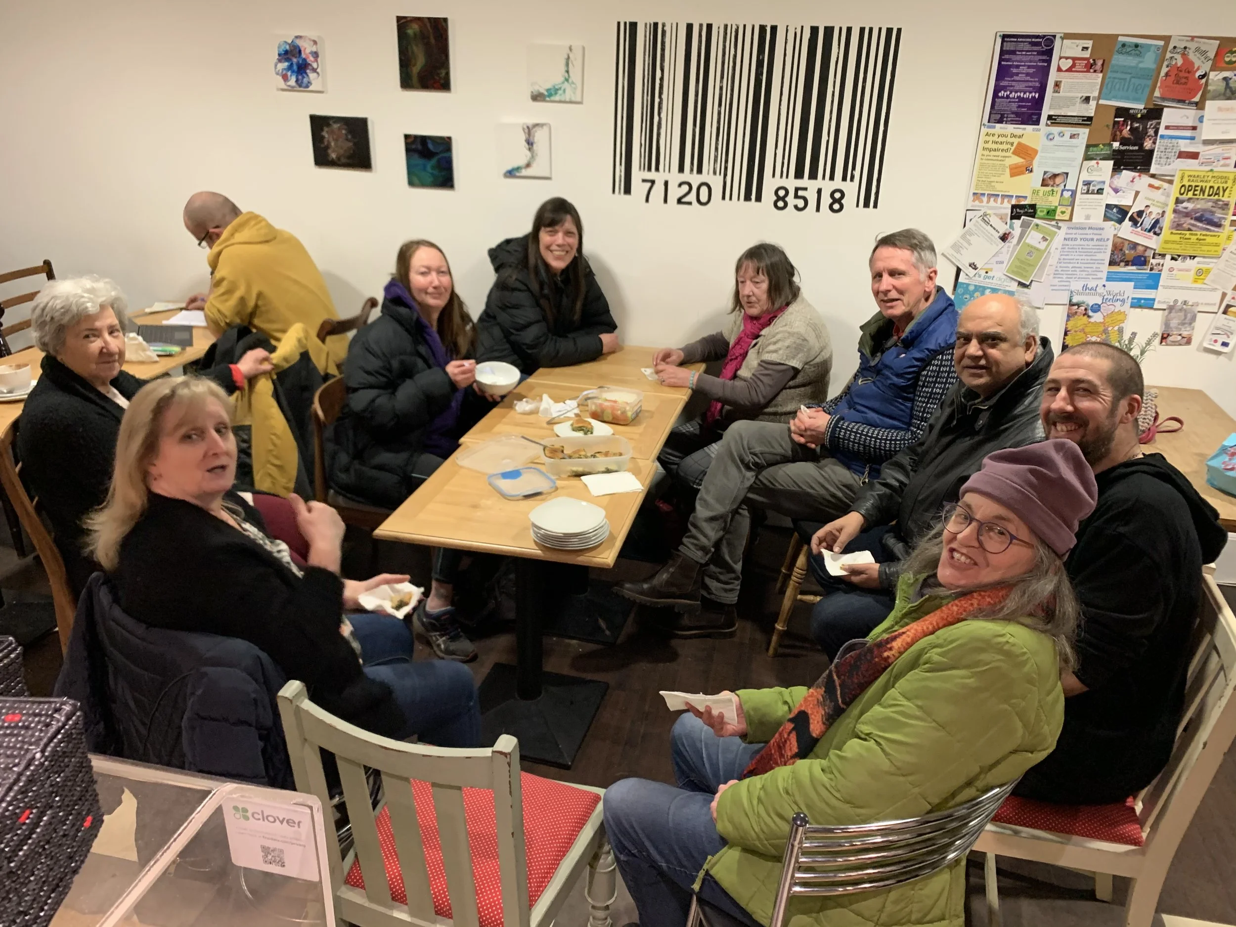 Group of people sitting around a rectangular wooden table enjoying a meal, with various dishes and bowls on the table in a room decorated with abstract art, a large barcode mural, and a bulletin board with flyers and posters.
