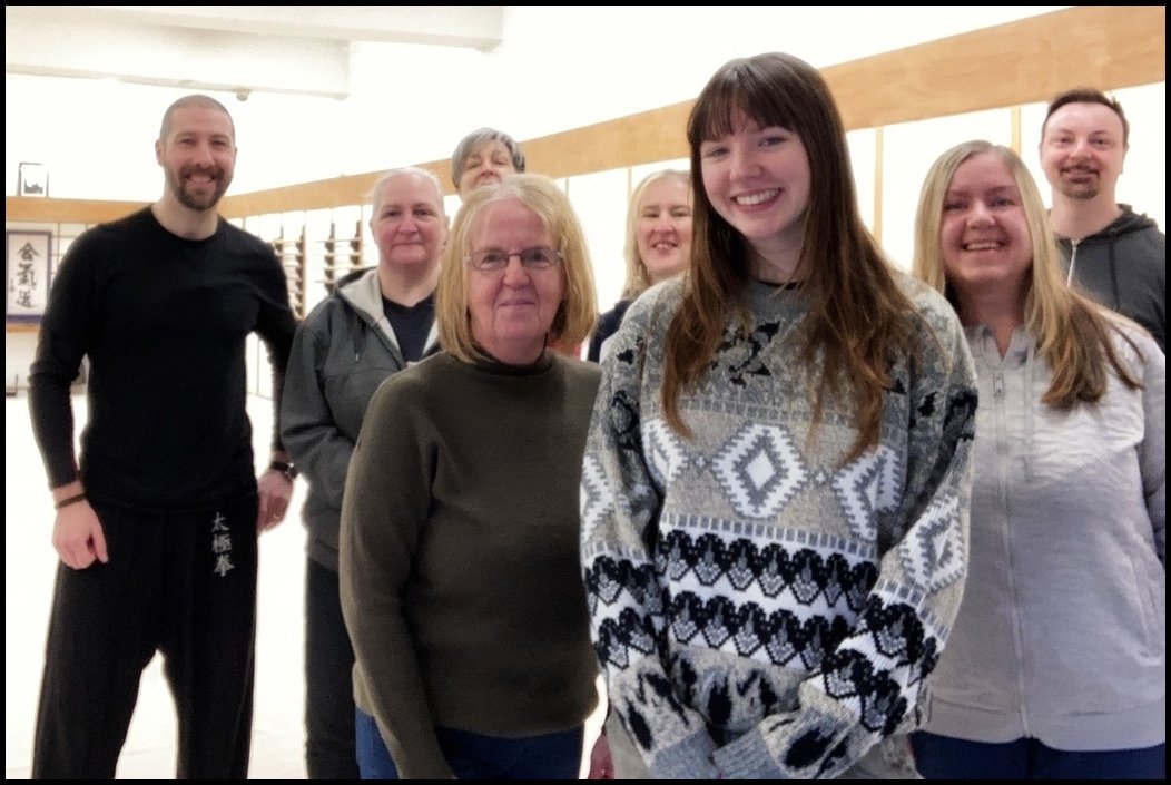 Group of seven smiling people standing in a row indoors, some wearing casual clothing, with wooden cabinets and framed artworks on the wall behind them.