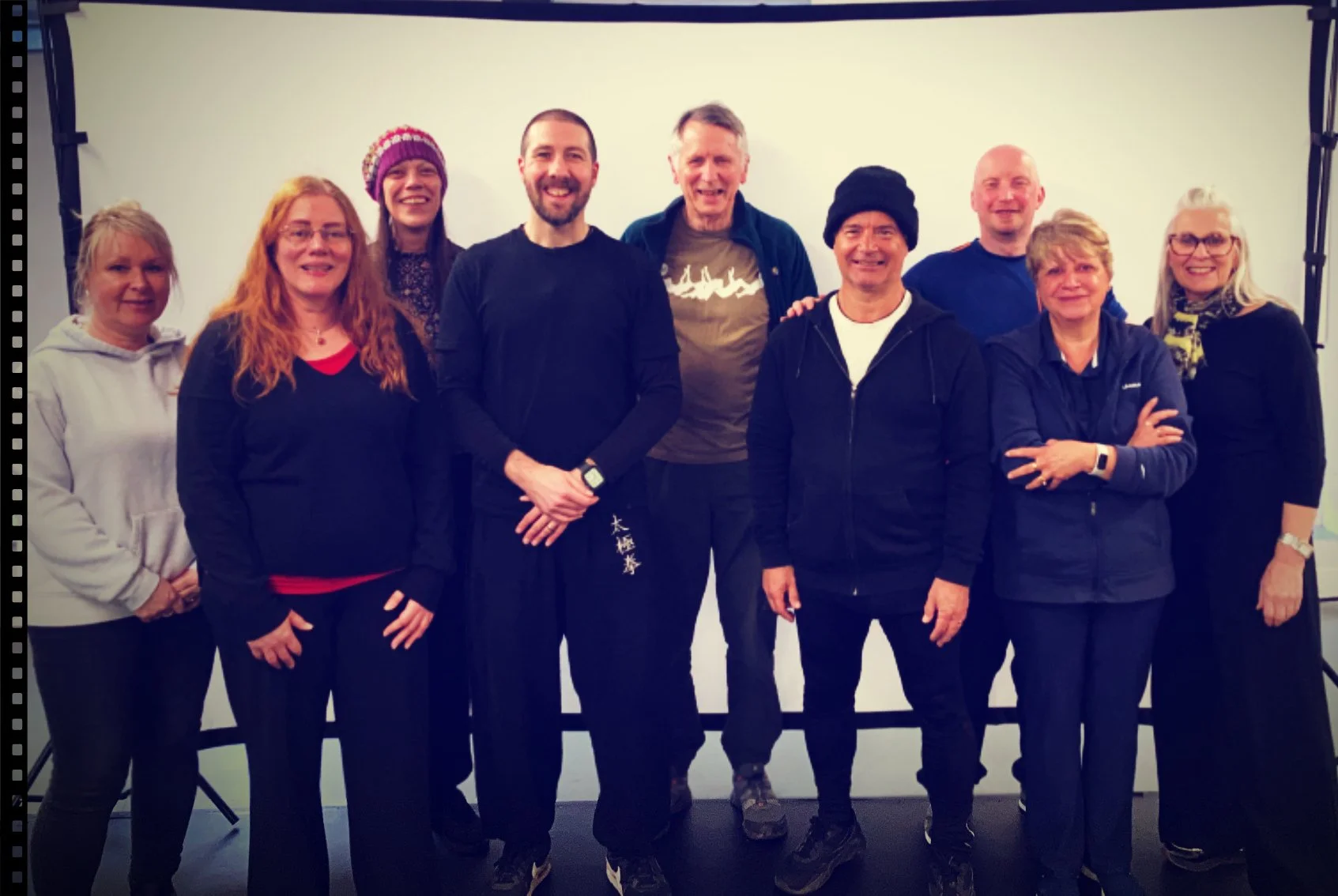 Group of ten Qigong participants standing together indoors in front of a plain white backdrop, smiling at the camera.