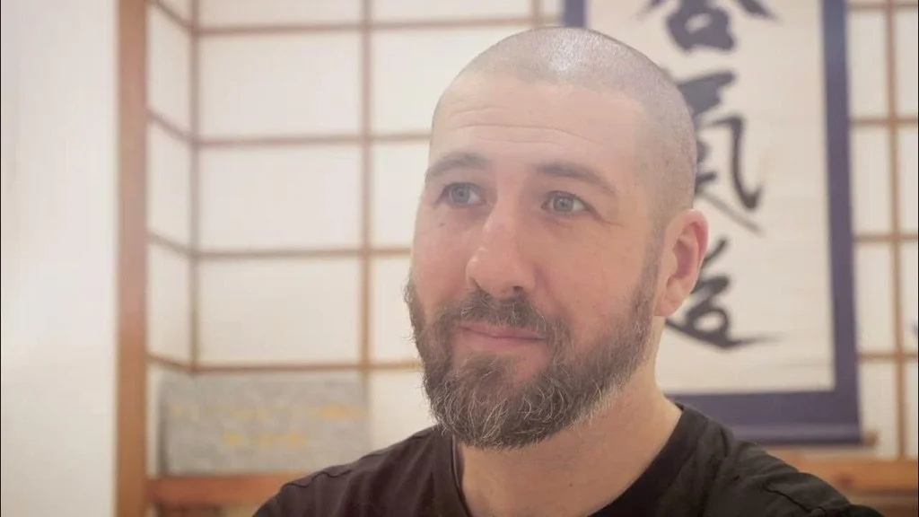 A man with a shaved head and beard looking to the side, sitting in front of a Japanese-style shoji screen with calligraphy artwork.
