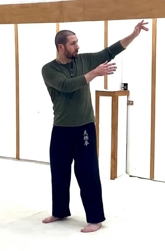 A man practicing martial arts in a dojo, wearing black martial arts pants with Japanese characters and a green long-sleeve shirt, demonstrating a move with one arm extended upwards and the other across his chest.