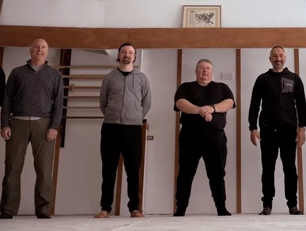 Qigong group standing indoors in front of a wooden frame and white wall, with a framed picture above them, posing for a group photo.