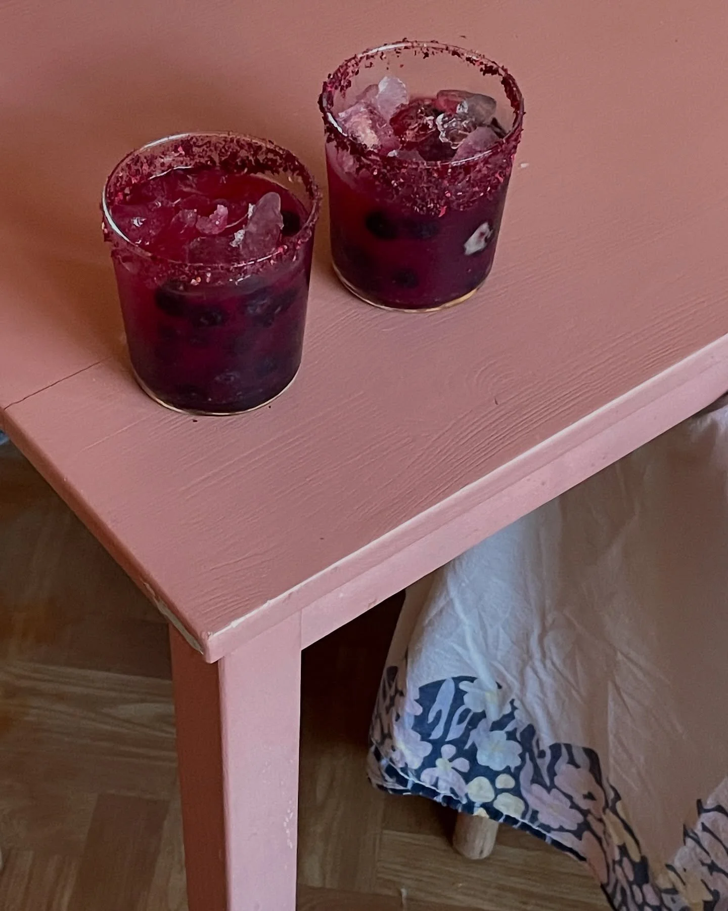 Two glasses filled with dark purple beverages, ice, and berries on a light pink wooden table. Part of a plastic bag with a black and white pattern is visible underneath the table.