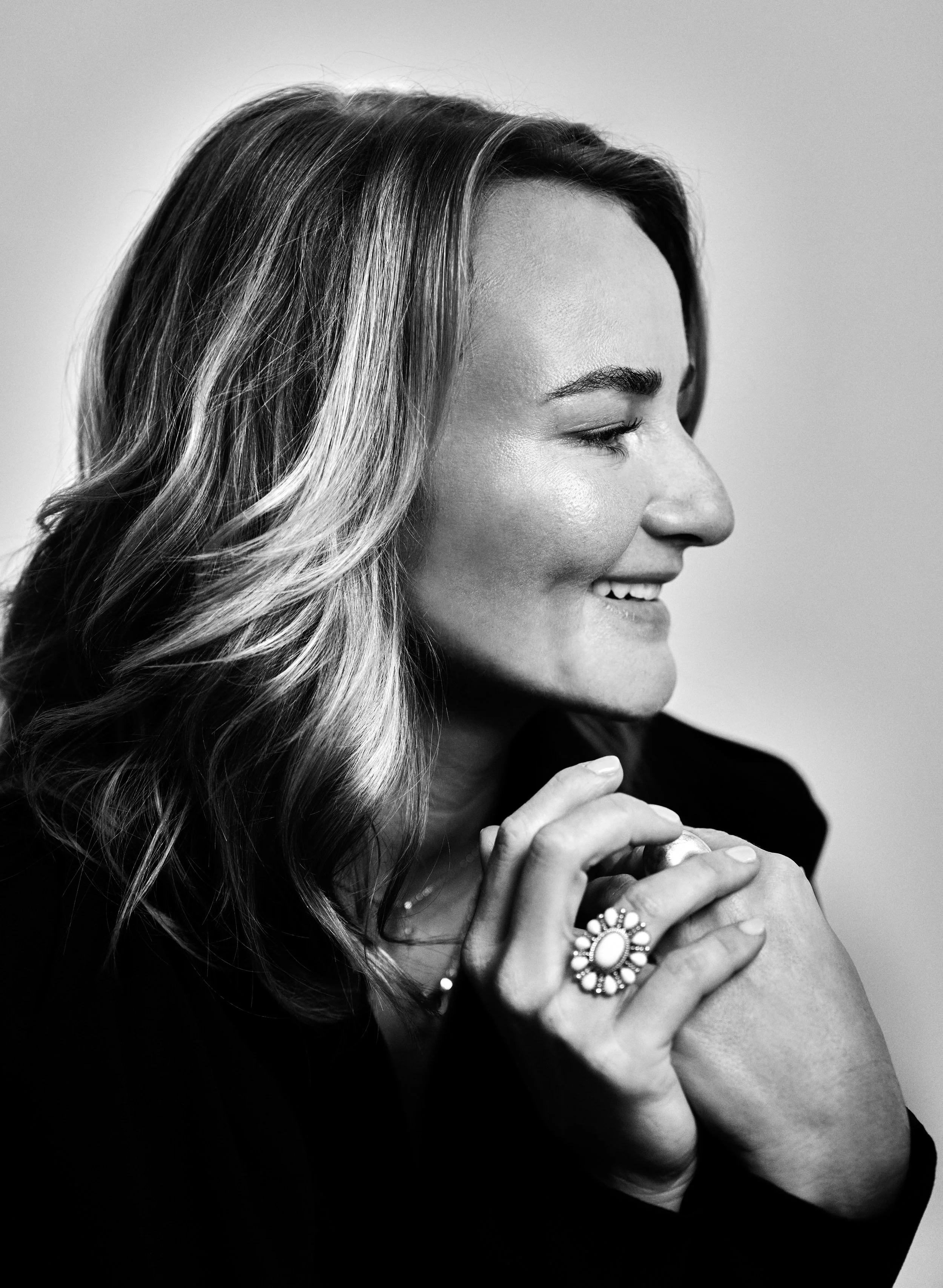 Zwart-wit foto van een glimlachende vrouw met golvend haar die haar handen onder haar Kin vouwt, met grote sieraden.