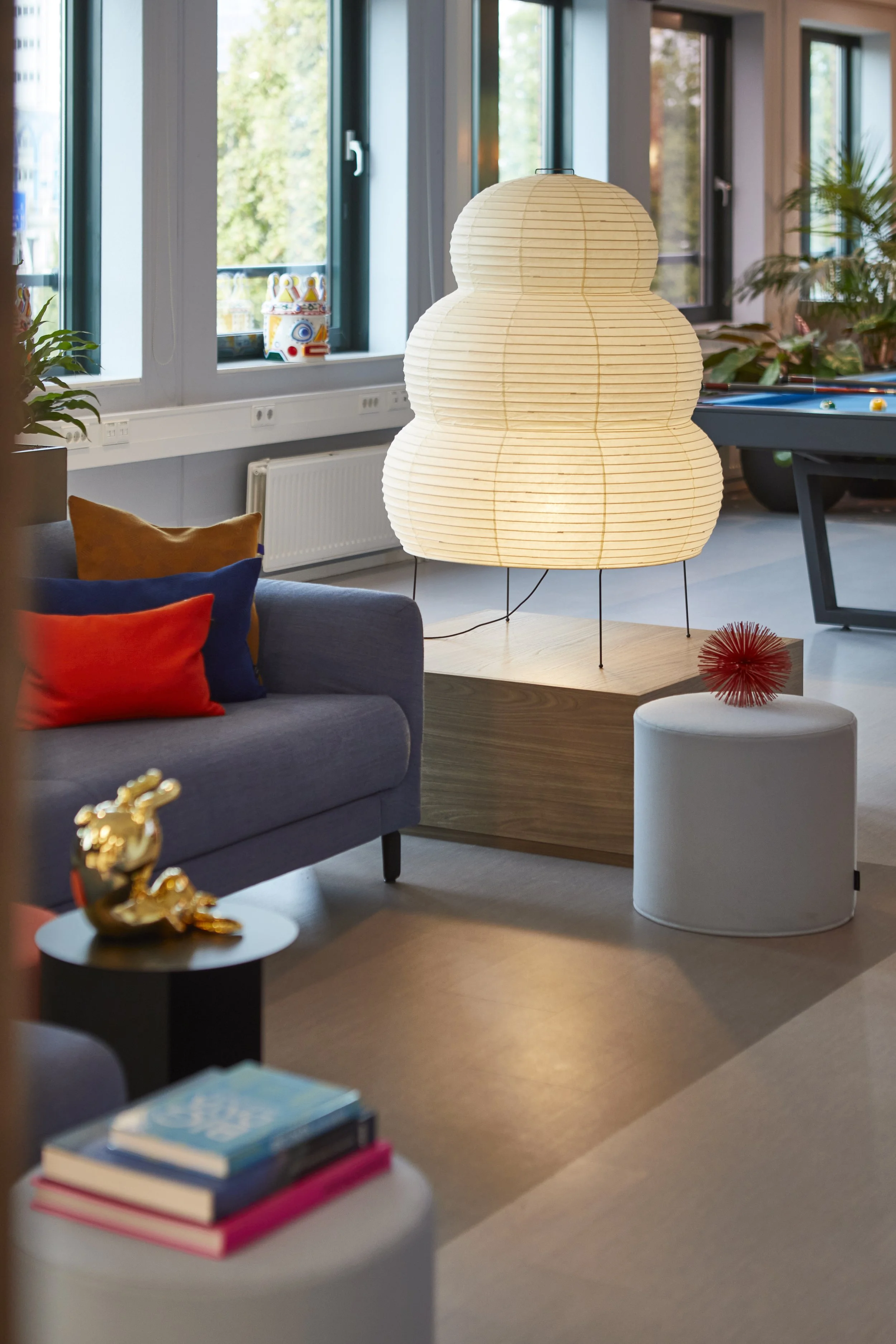 Interieur van een woonkamer met een grote papieren lamp, een bank met kleurrijke kussens, een boekenkast en een biljarttafel op de achtergrond.