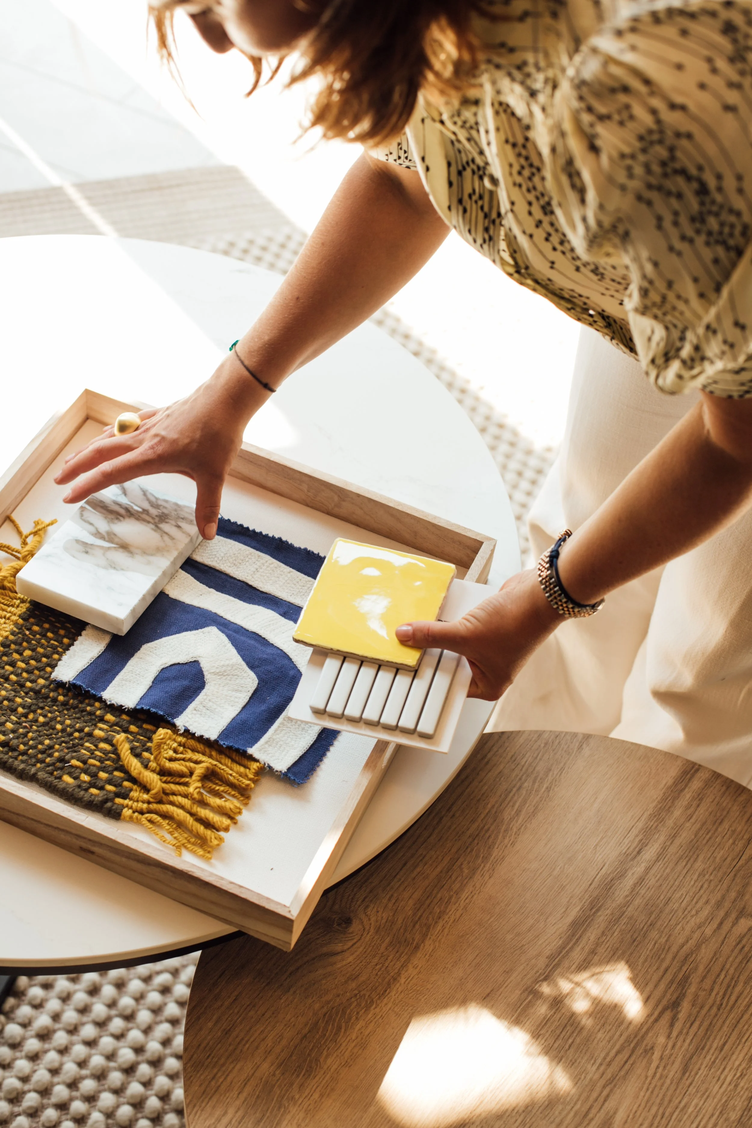 Een persoon plaatst decoratieve items op een witte tafel, met een tray met stoffen, tegels en een stukje geborsteld geel materiaal.