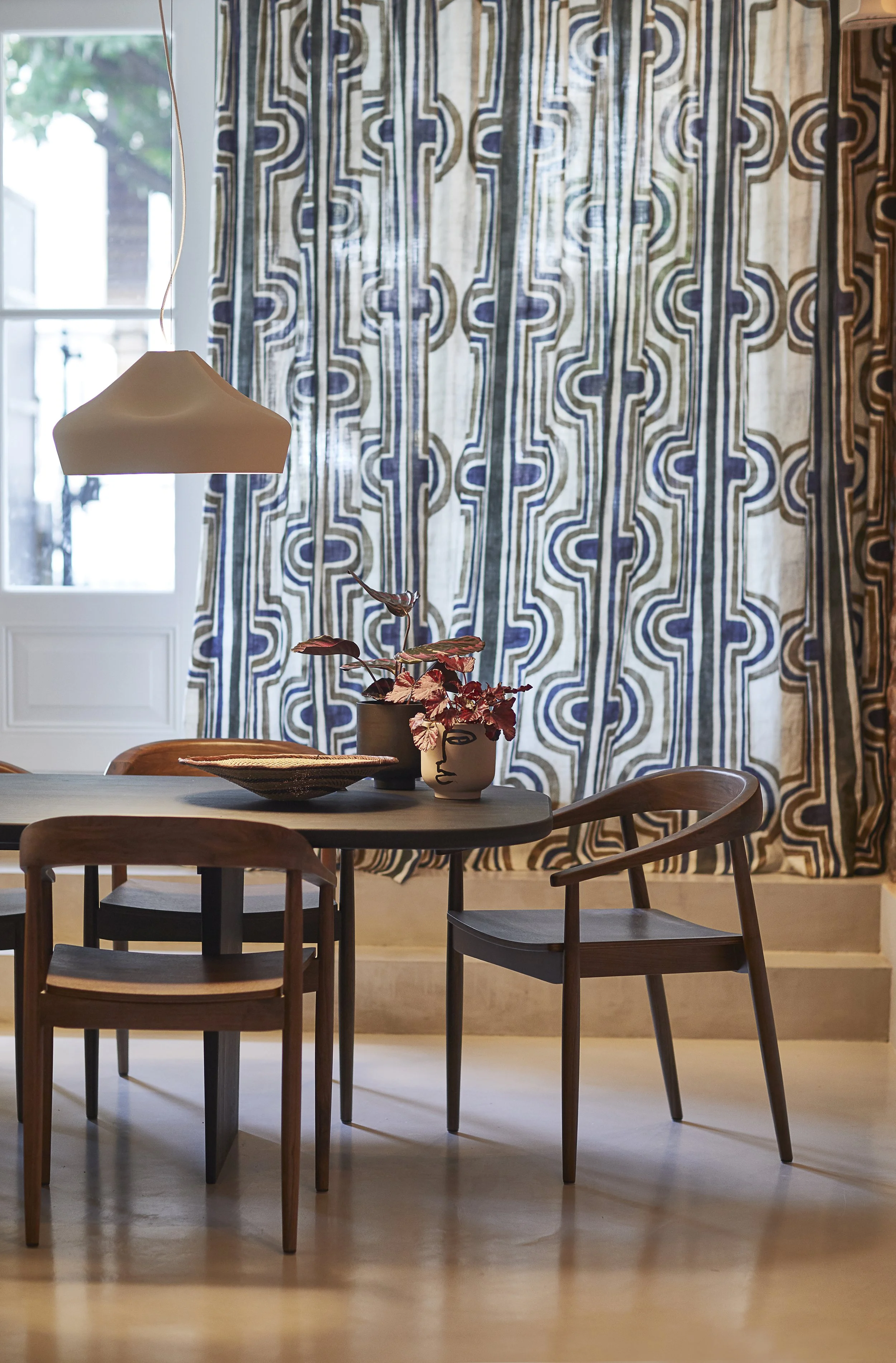 Binnenruimte met een eettafel, drie houten stoelen, een hanglamp en een mooie gordijn met blauwe en beige patronen.