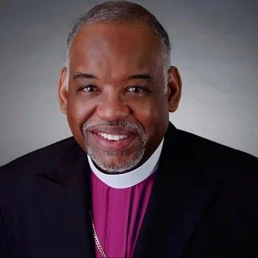 Portrait of a smiling man in clergy attire with a purple clerical shirt, black jacket, and a white collar.