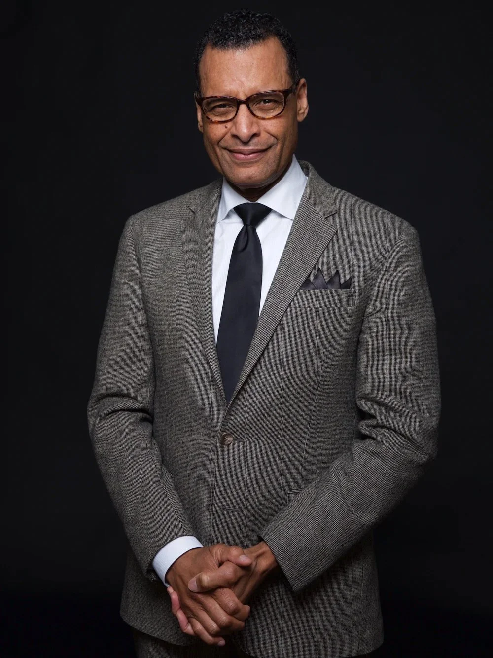 A man in a gray suit with a white shirt and black tie, wearing glasses, standing against a black background.