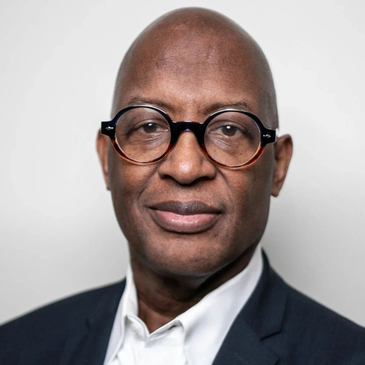 A middle-aged man wearing glasses, a dark suit, and a white shirt, looking directly at the camera with a neutral expression.