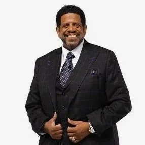 Smiling man in a dark suit with a checkered shirt and tie, standing against a white background.