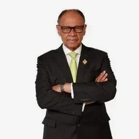 An elderly man in a black suit with a green tie and glasses, standing confidently with arms crossed against a white background.