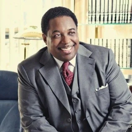 A man in a gray suit and pink tie smiling, sitting in a well-lit office or library with shelves of books in the background.