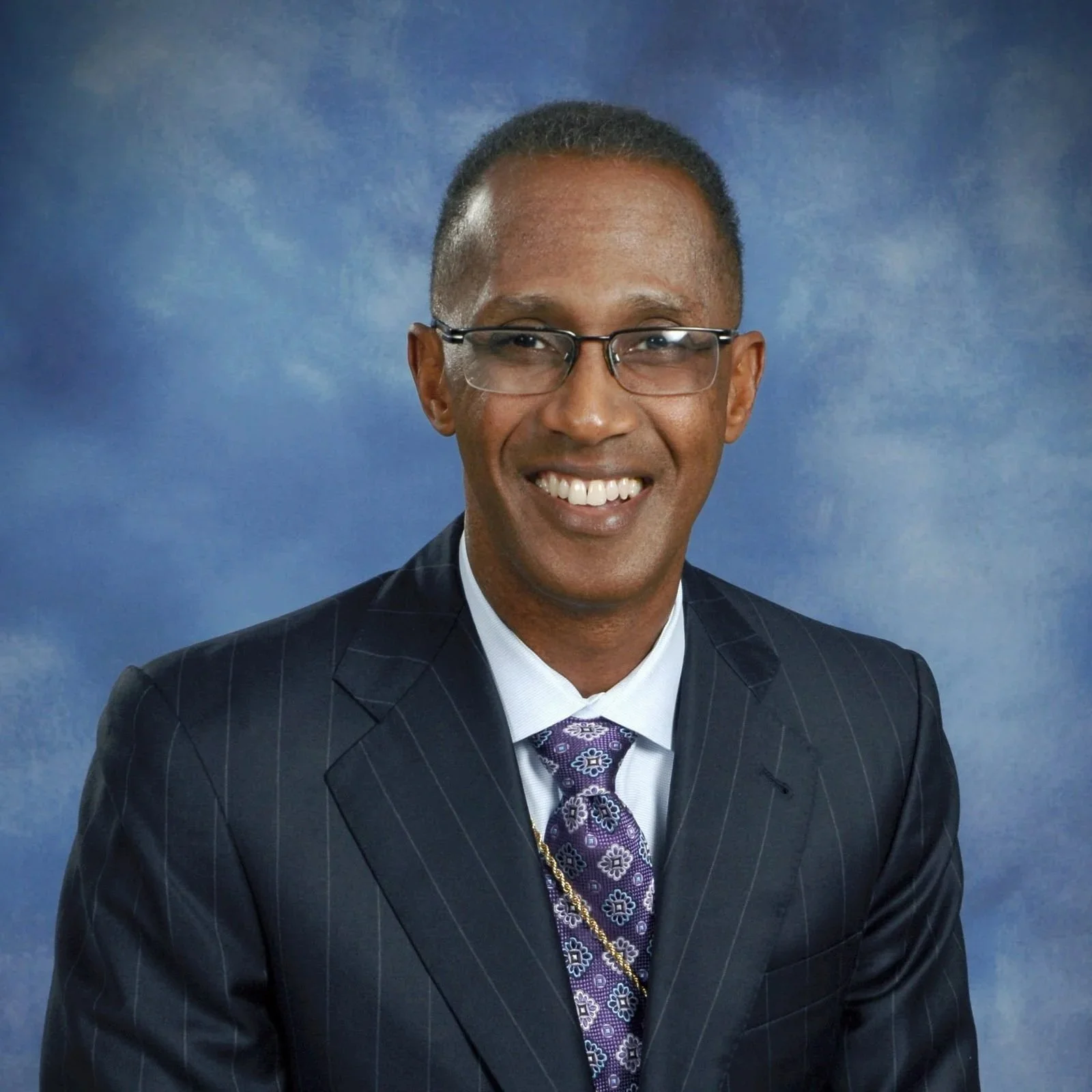 A smiling man wearing glasses, a dark pinstripe suit, a white shirt, and a purple patterned tie, posed against a blue background.