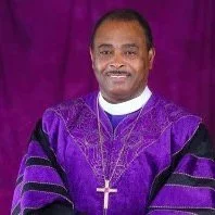 Photograph of a man in purple clerical robes with a cross necklace standing in front of a purple curtain.