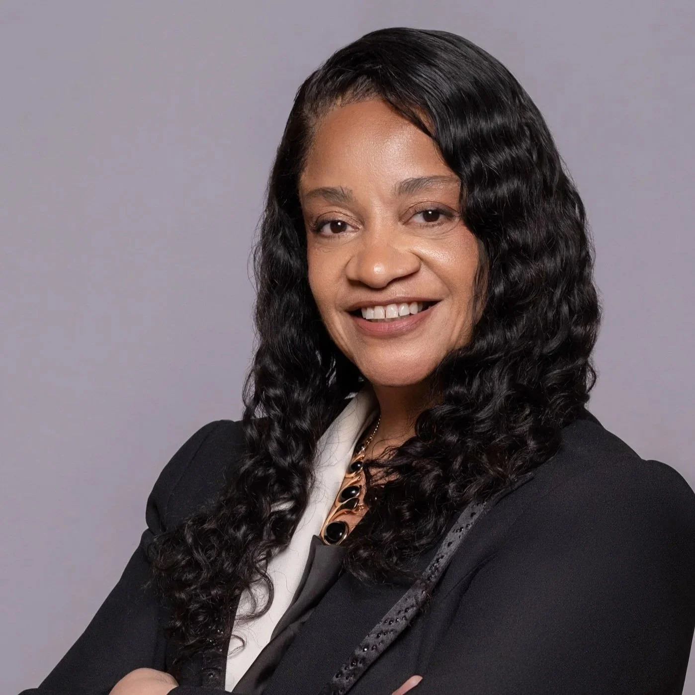 Portrait of an African American woman with curly black hair, wearing a dark blazer and blue top, smiling, with a bookshelf in the background.