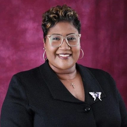A woman with short curly hair, wearing glasses, hoop earrings, and a black blazer with a butterfly pin, smiling against a purple background.