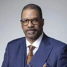 A professional man wearing a blue suit, white shirt, and striped tie, with glasses and a serious expression.