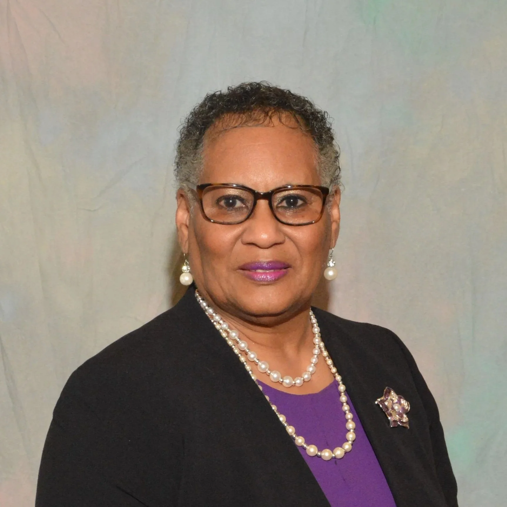 A woman with short, curly gray hair wearing glasses, pearl earrings, a pearl necklace, a purple dress, and a black blazer. She has pink lipstick and a decorative brooch on her blazer.