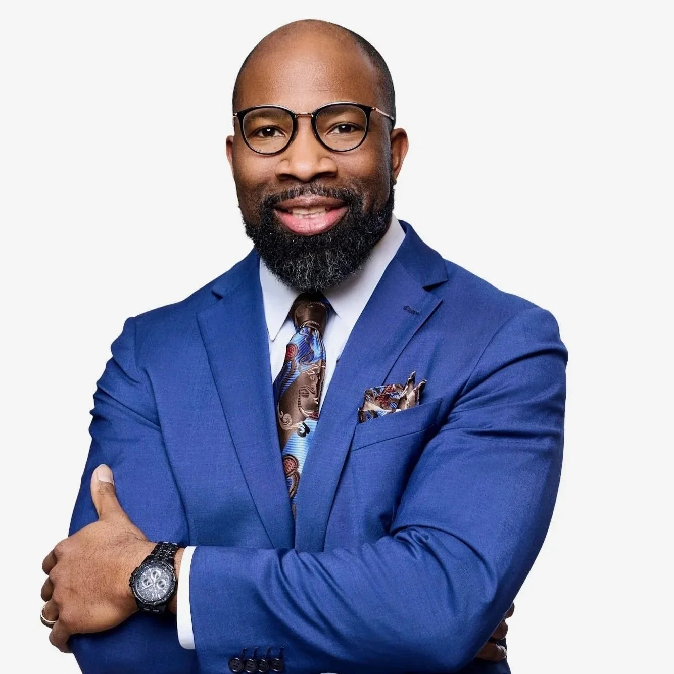 Portrait of a man in a blue suit, wearing glasses, with a beard, smiling, against a white background.