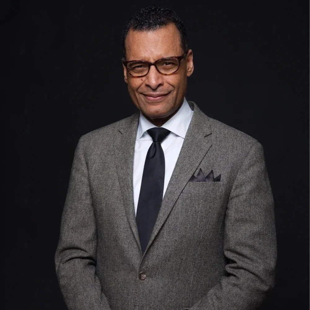 A man in a grey suit, white shirt, black tie, and glasses smiling against a black background.