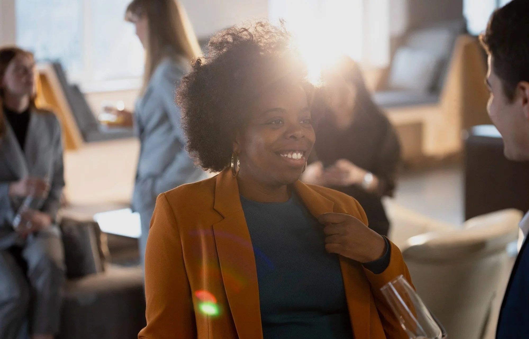 A woman with an afro hairstyle smiling at a networking event, wearing an orange blazer and holding a glass of wine, engaging in conversation with a man.