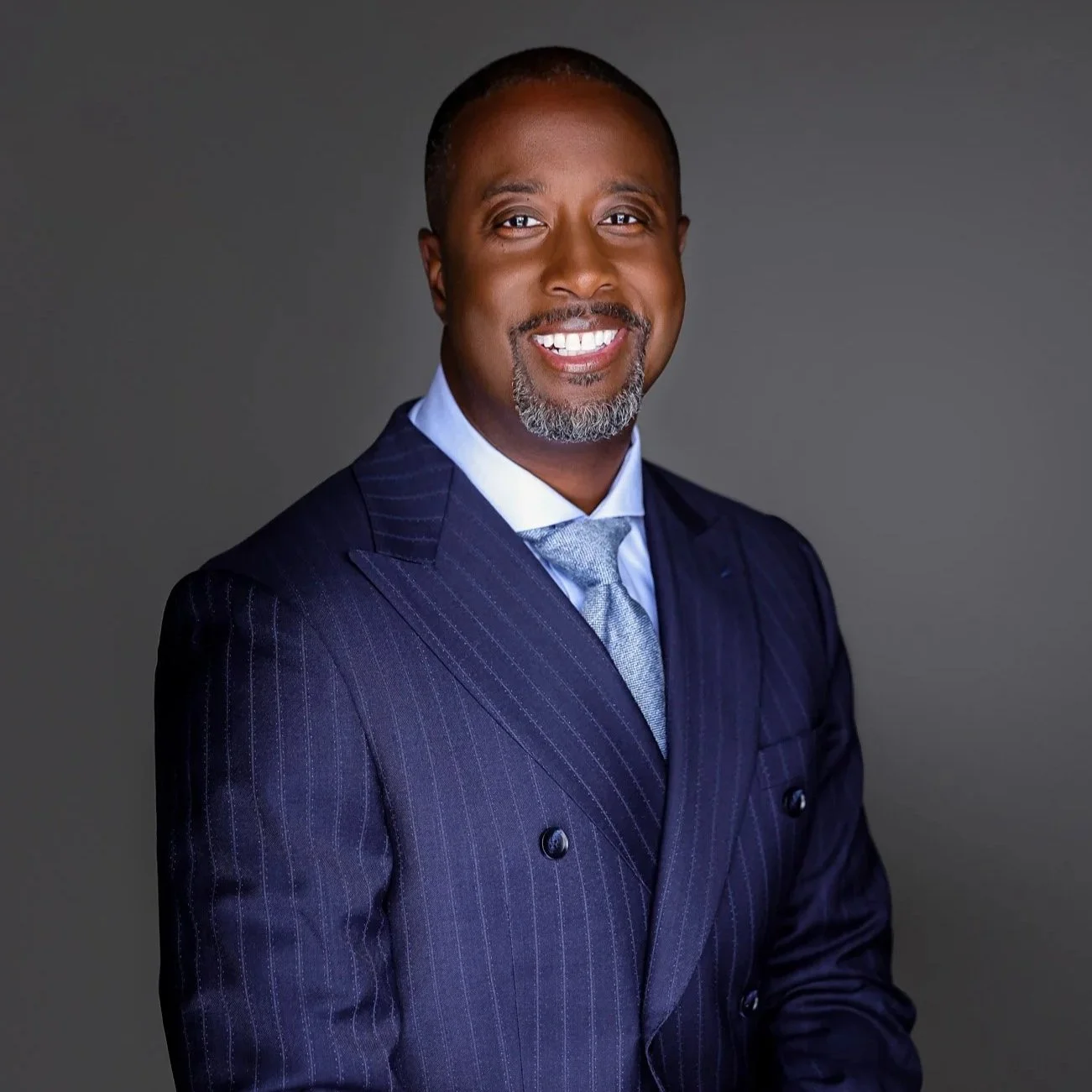 A professional African American man smiling in a dark blue pinstripe suit with a light blue shirt and tie, against a plain gray background.