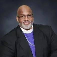 Portrait of an older African American man with glasses, beard, wearing a black suit jacket, purple shirt, and a chain necklace, against a dark background.