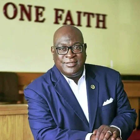 A man wearing glasses and a blue suit, sitting at a table with a serious expression, with a sign that says "ONE FAITH" in the background.