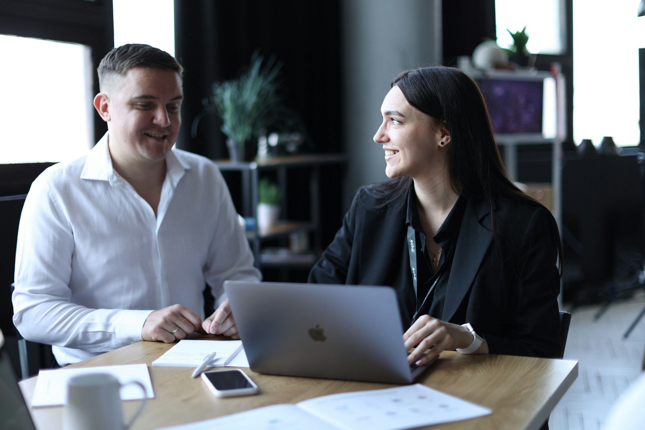 Two people, a man and a woman, talking and smiling at a meeting table with a laptop, phone, mug, and papers in a modern office.