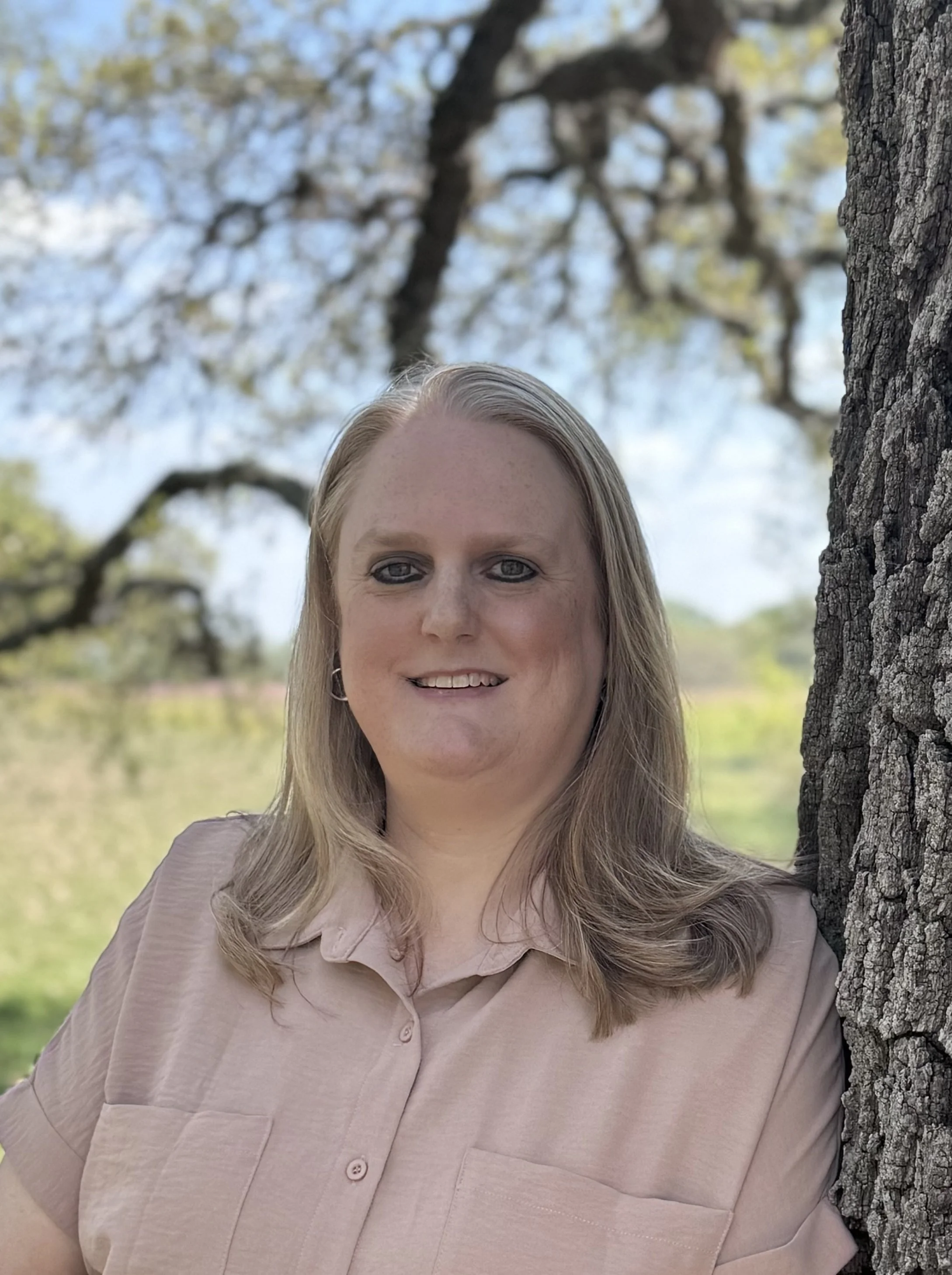 A woman with blonde hair, smiling slightly, standing outdoors beside a large tree trunk with a blurred background of trees and sky.