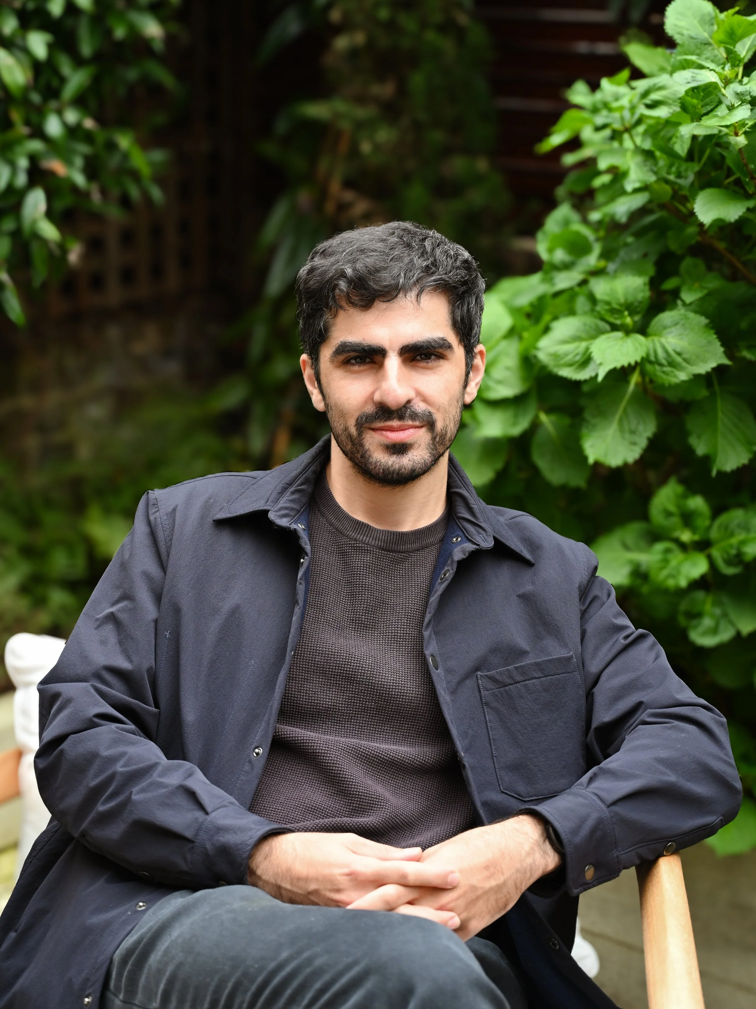 A man with dark hair and a beard sitting outdoors in front of green leafy plants, wearing a dark jacket and a gray shirt, with a neutral expression.